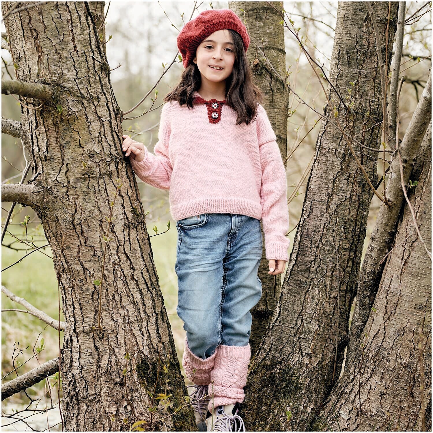 Essentials Super aran: Mädchen in rosa Pullover von Rico Design, steht zwischen Bäumen, trägt Jeans.