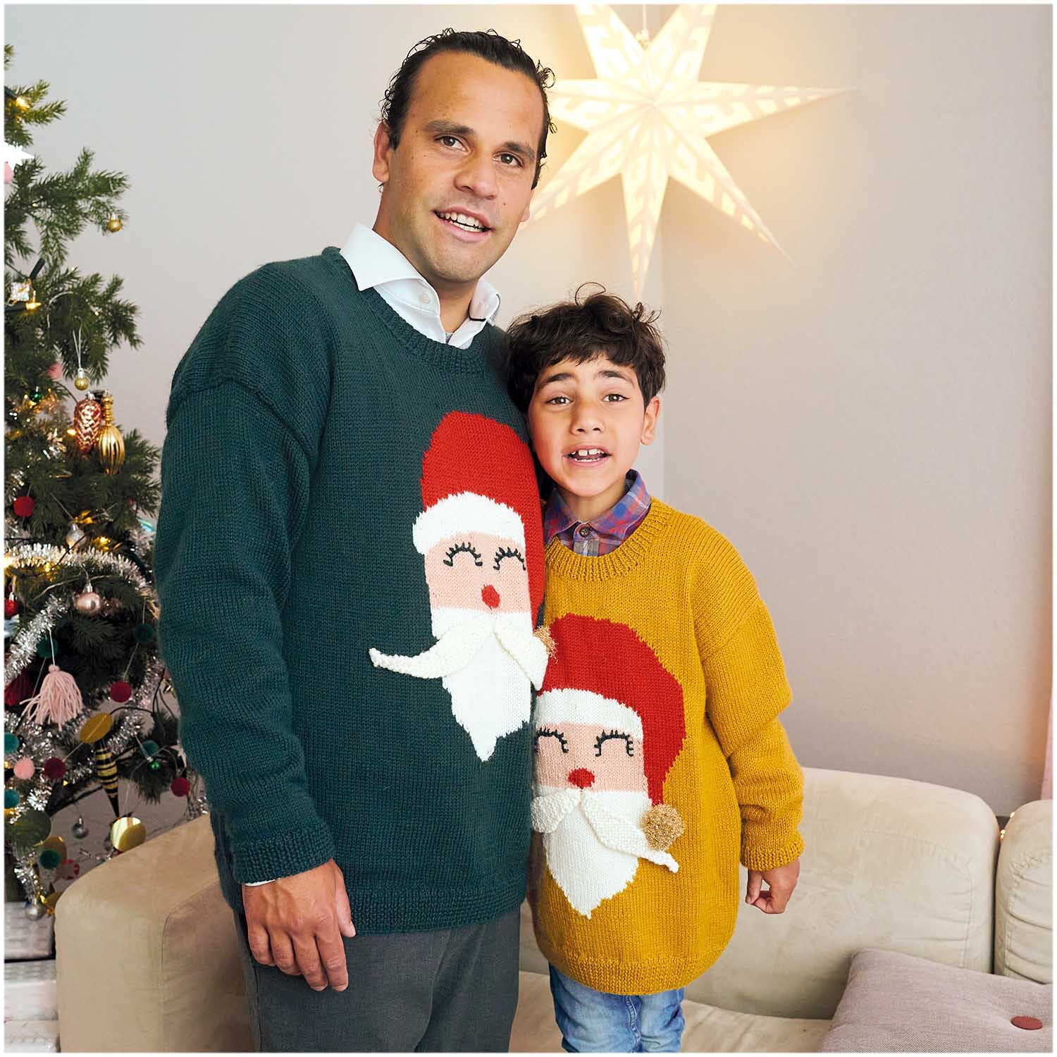 Essentials Merino Plus dk: Vater und Sohn tragen weihnachtliche Pullover mit Santa-Motiv vor einem festlich geschmückten Baum.