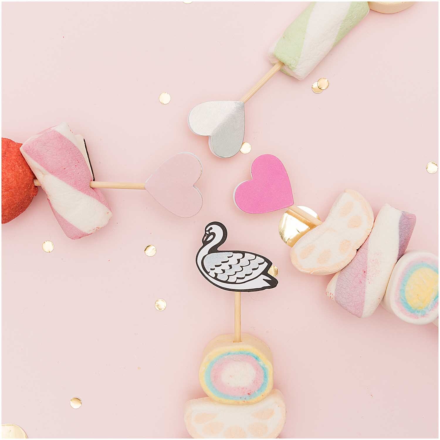 Picker Mix Schwan irisierend 15cm 24 Stück: Bunte Marshmallow-Spieße mit Schwan-Design auf rosa Hintergrund, dekoriert mit Herzen.