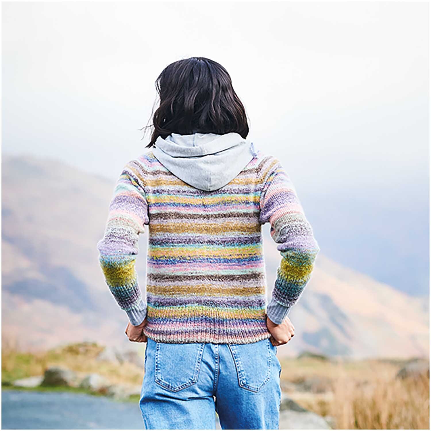 Strickset Pullover Modell 01: Frau in gestreiftem Pullover und Jeans vor Berglandschaft von hinten.
