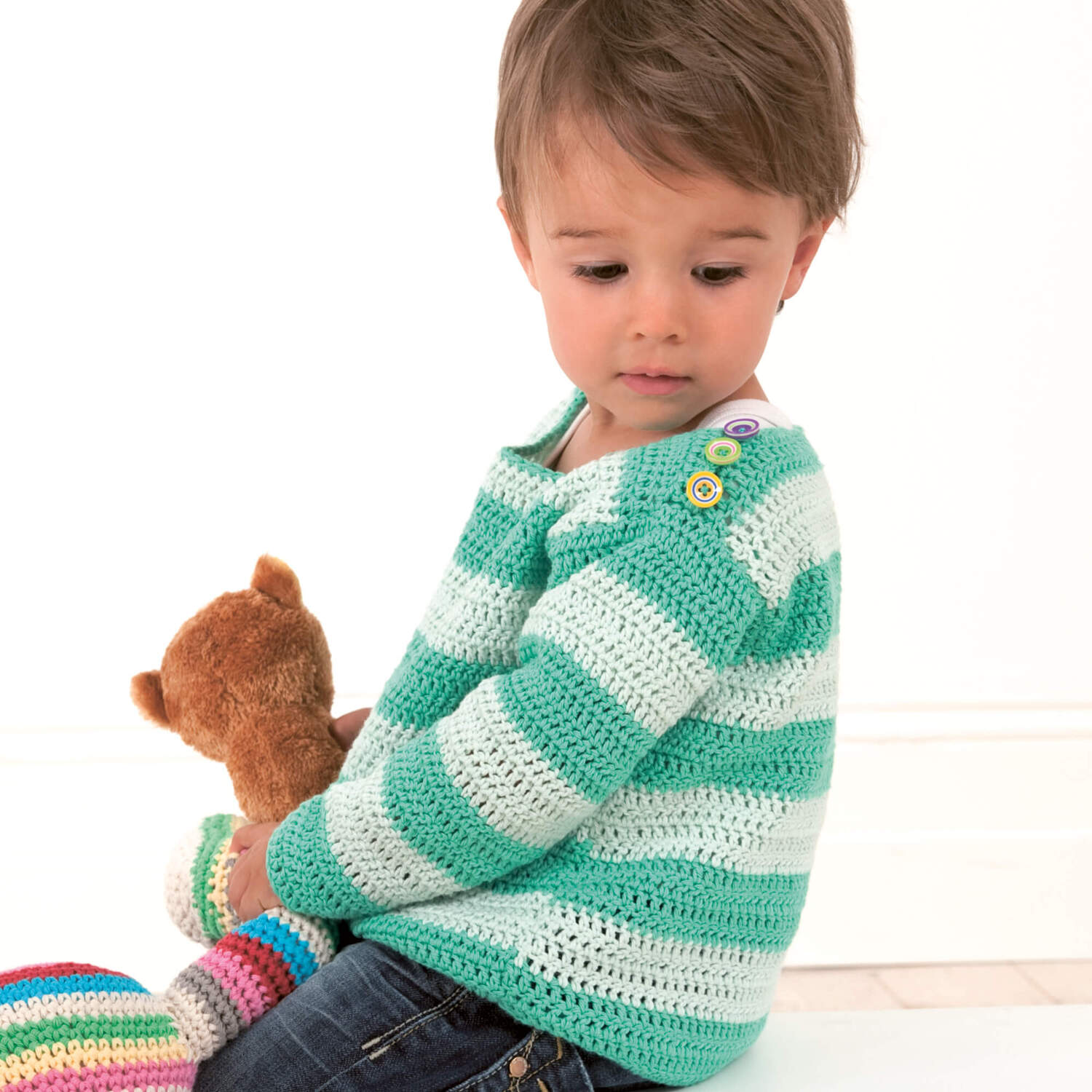 Häkelanleitung Kinder-Streifenpullover: Kind trägt türkis-weiß gestreiften Pullover mit Knöpfen, hält ein Kuscheltier.