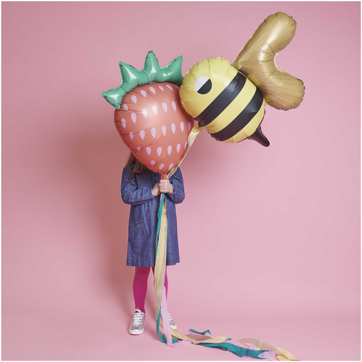 Folienballon Biene 60x70cm von Rico Design: Kind hält Bienen- und Erdbeerballon vor rosa Hintergrund.