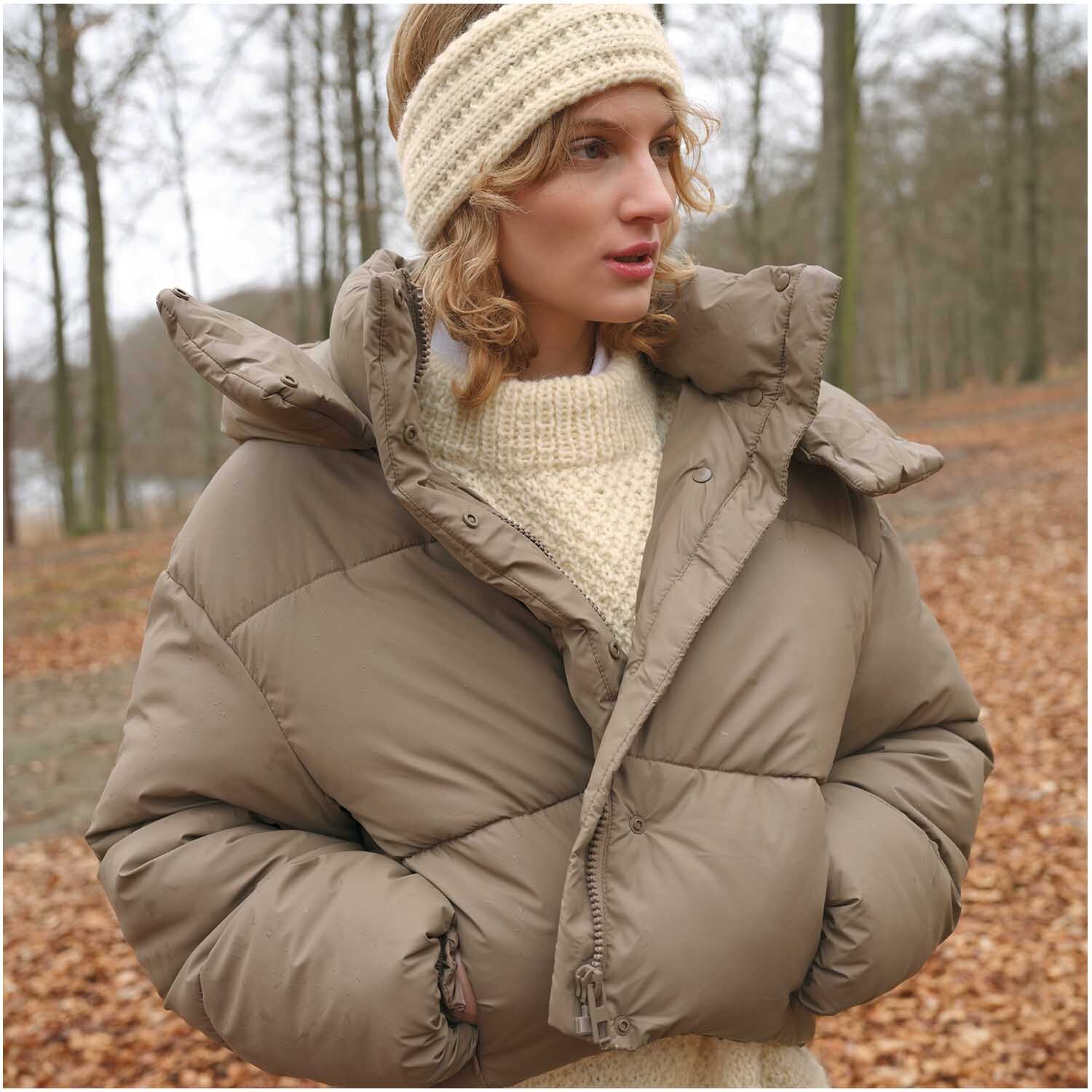 Strickset Stirnband Modell 12: Frau im Wald mit beigem Stirnband und dicker, brauner Winterjacke.