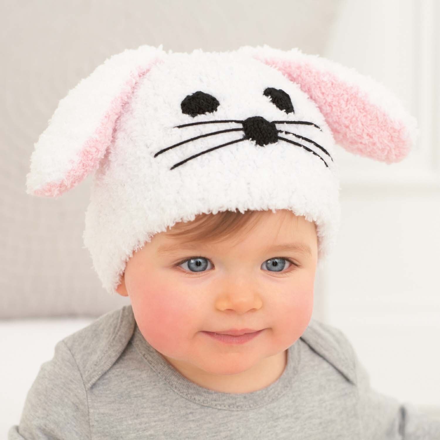 Strickanleitung Babymütze mit Hasenohren: Baby trägt weiße Mütze mit rosa Ohren und Hasengesicht.