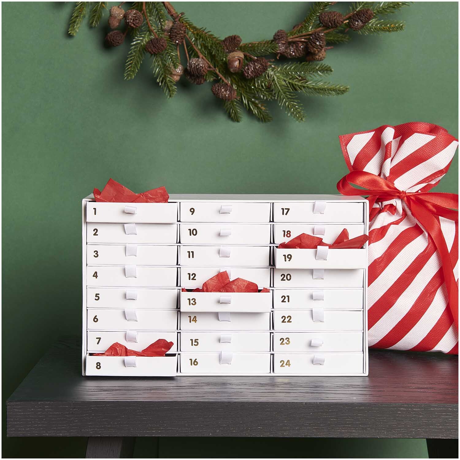 Adventskalender Box weiß-gold von Rico Design auf einem Tisch, dekoriert mit Tannenzweigen und einem gestreiften Geschenkbeutel.