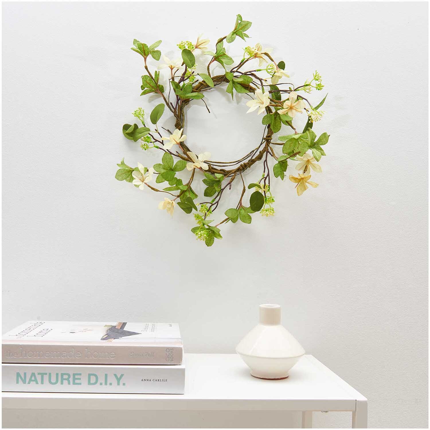 Kranz mit Blüten creme-weiß 40cm von Rico Design, dekorativ an einer weißen Wand über einem Tisch mit Büchern und Vase.