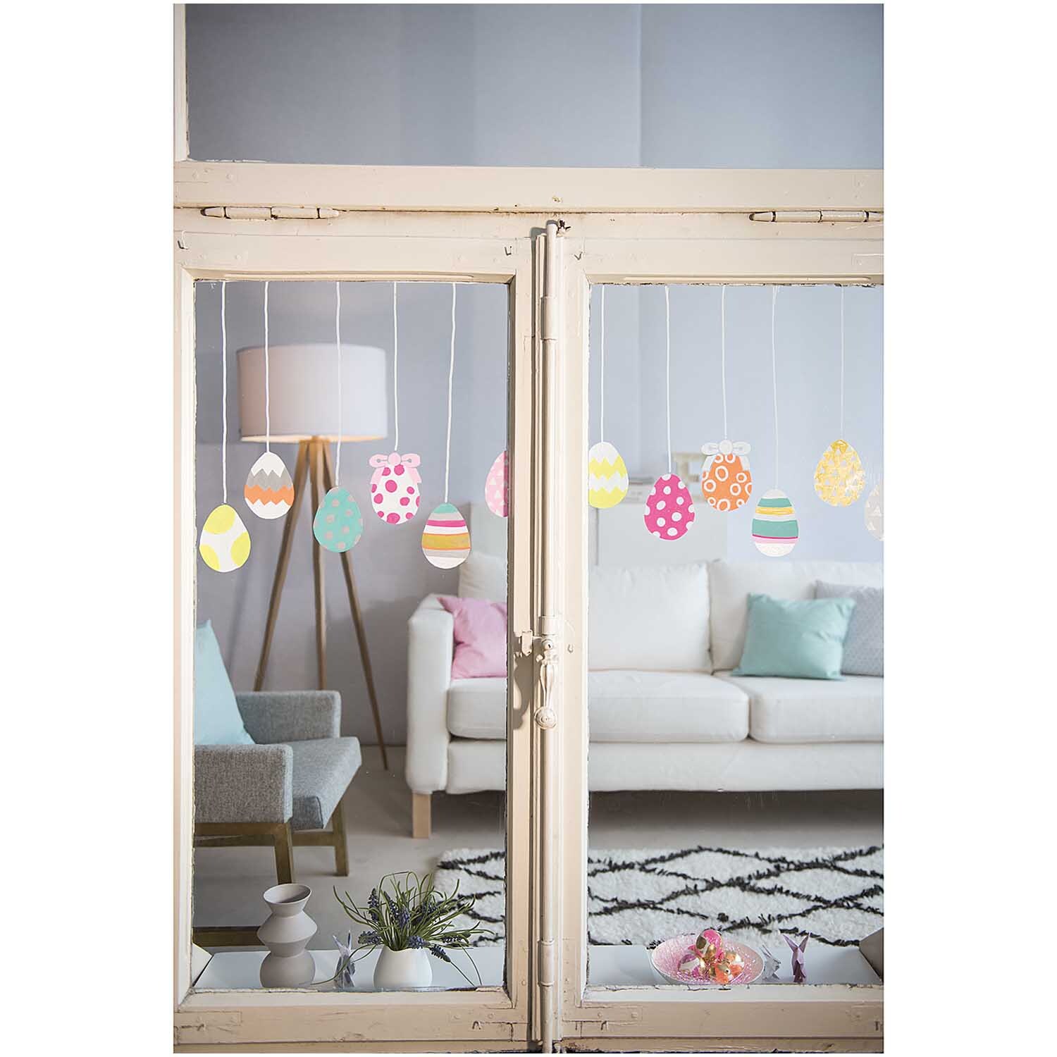 Vorlagenmappe Ostern Fenster- und Glasmalerei mit bunten Eiern, dekoriert mit Kreidestiften, im Wohnzimmer mit Sofa und Kissen.