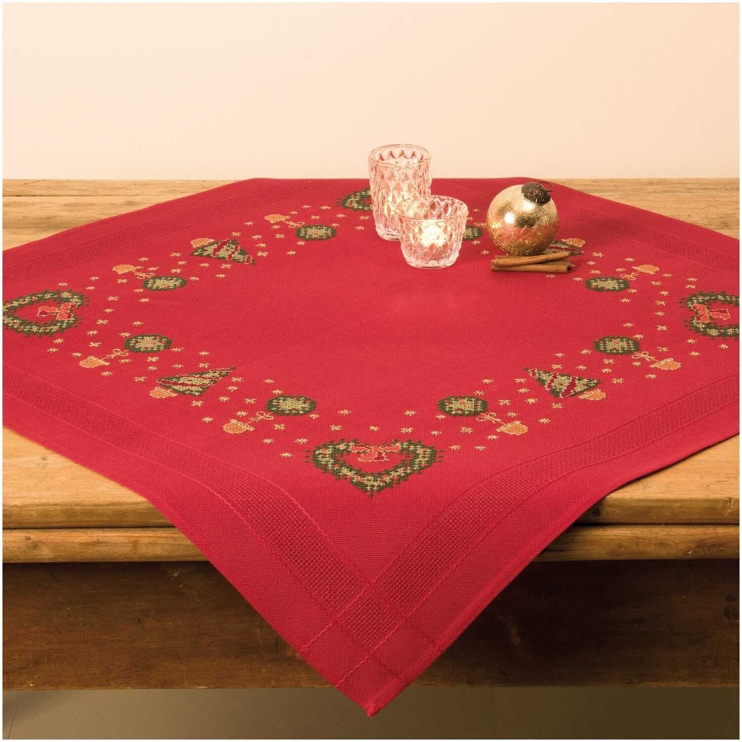 Stickpackung Tischdecke Buchsbaumherz 80x80cm zeigt ein rotes Design mit grünen Herzmotiven, dekoriert mit Kerzen und Kugeln auf einem Holztisch.
