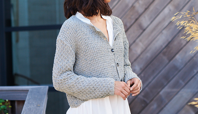 Strickanleitung Strukturmuster-Jacke aus Fashion Cotton Neps dk: Frau trägt graue Strickjacke über weißem Kleid vor Holzwand.