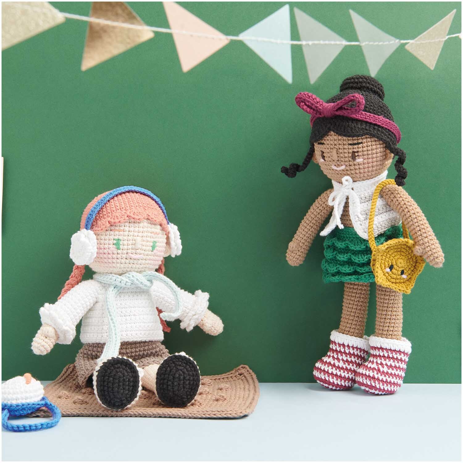 Creative Ricorumi dk: Zwei gehäkelte Puppen vor grünem Hintergrund, eine sitzend mit Kopfhörern, die andere stehend mit Tasche.
