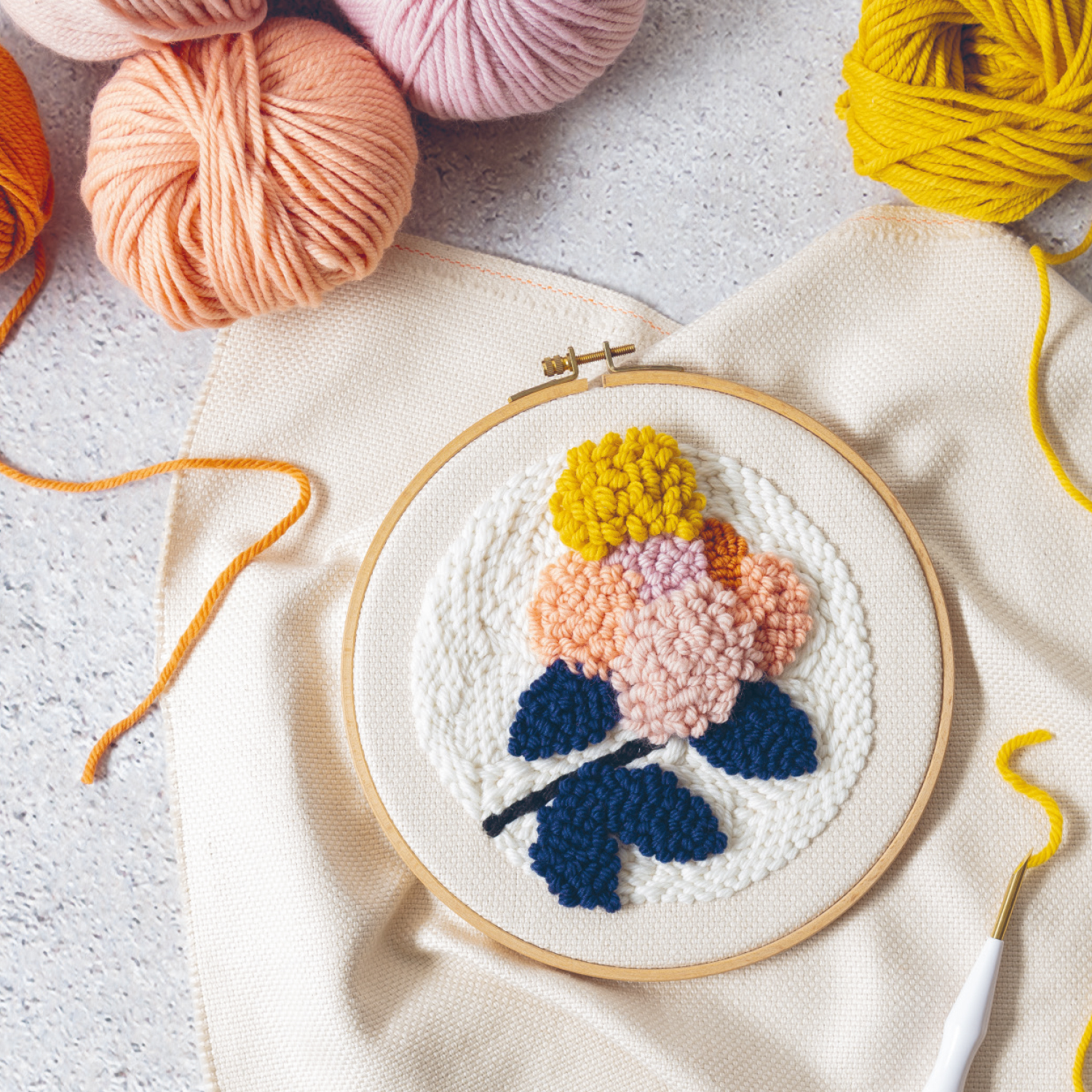 Punch Needle Anleitung Florale Punch Needle Deko mit buntem Blumenmotiv in Stickrahmen, umgeben von Garnknäueln in verschiedenen Farben.