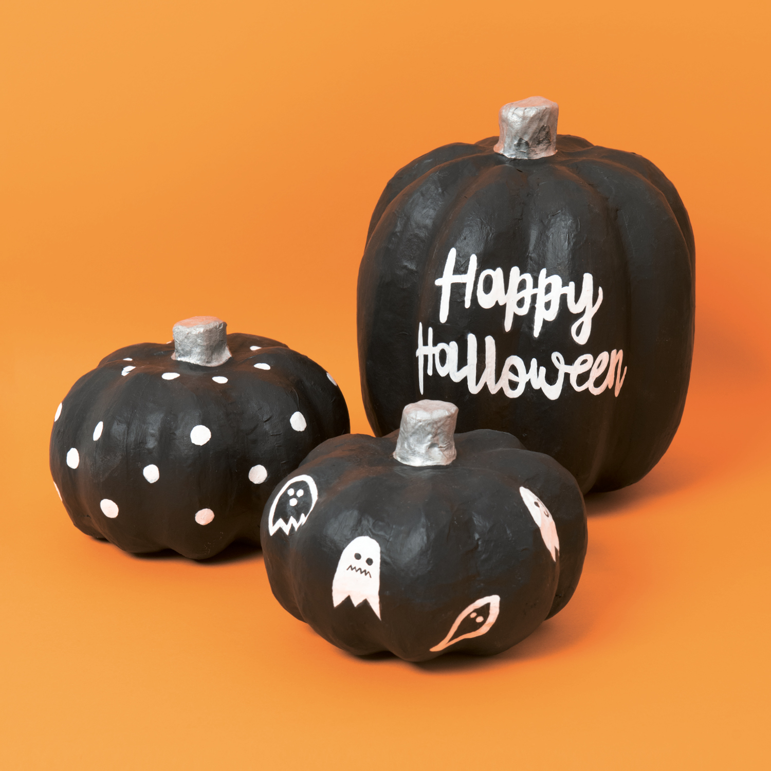 Anleitung Pappmaché-Kürbisse bemalen: Drei schwarze Kürbisse mit Halloween-Motiven auf orangefarbenem Hintergrund.