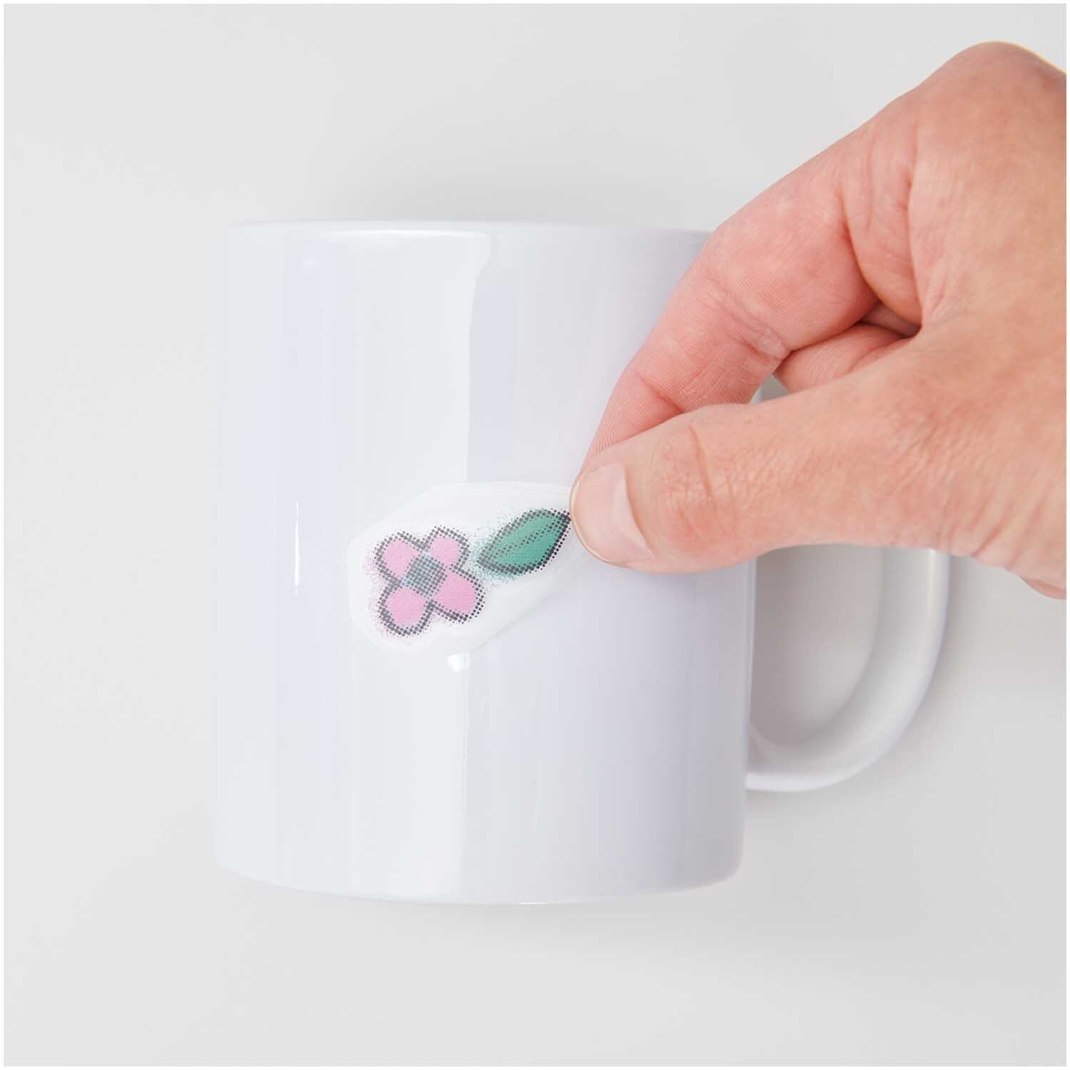 Transfermotiv Spray-Mix 10 Stück: Eine Hand klebt ein florales Motiv auf eine weiße Tasse. Marke: Rico Design.