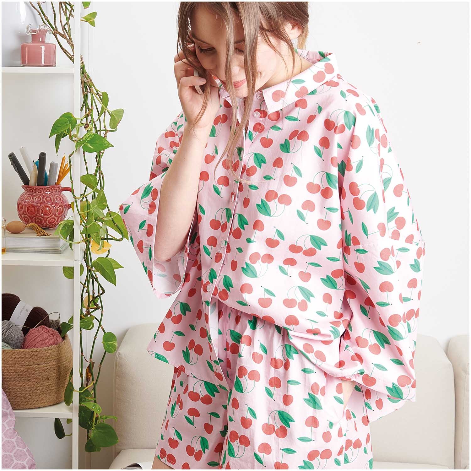 Meterware Baumwoll-Popelin rosa Kirschen: Frau in rosa Pyjama mit Kirschmuster, umgeben von Pflanzen und Garn. Ideal für kreative Nähprojekte.