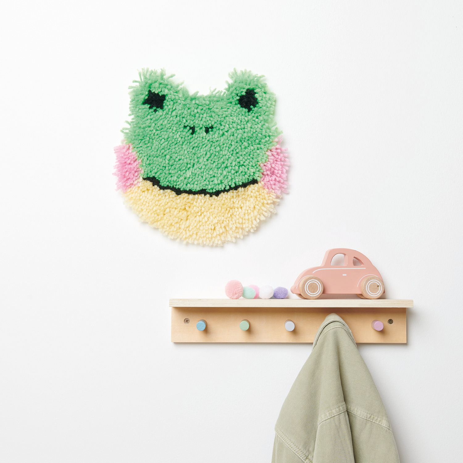 Rico Hooki Hooki Knüpfset Wandbehang Frosch zeigt einen bunten Frosch-Wandteppich über einer Garderobe.