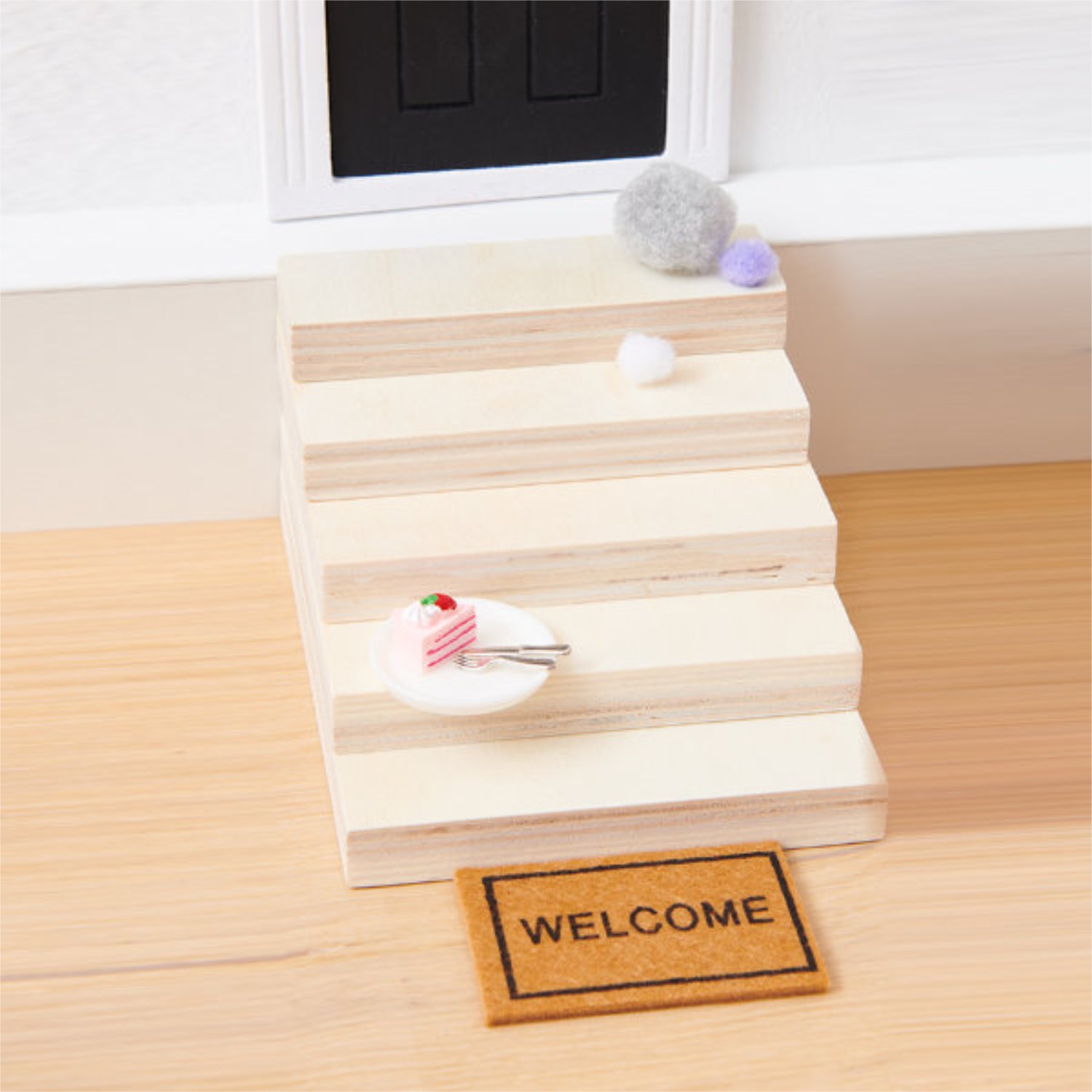 Miniatur-Holztreppe davor eine Fußmatte und auf der Treppe ein Miniatur-Teller mit einem Stück Torte.