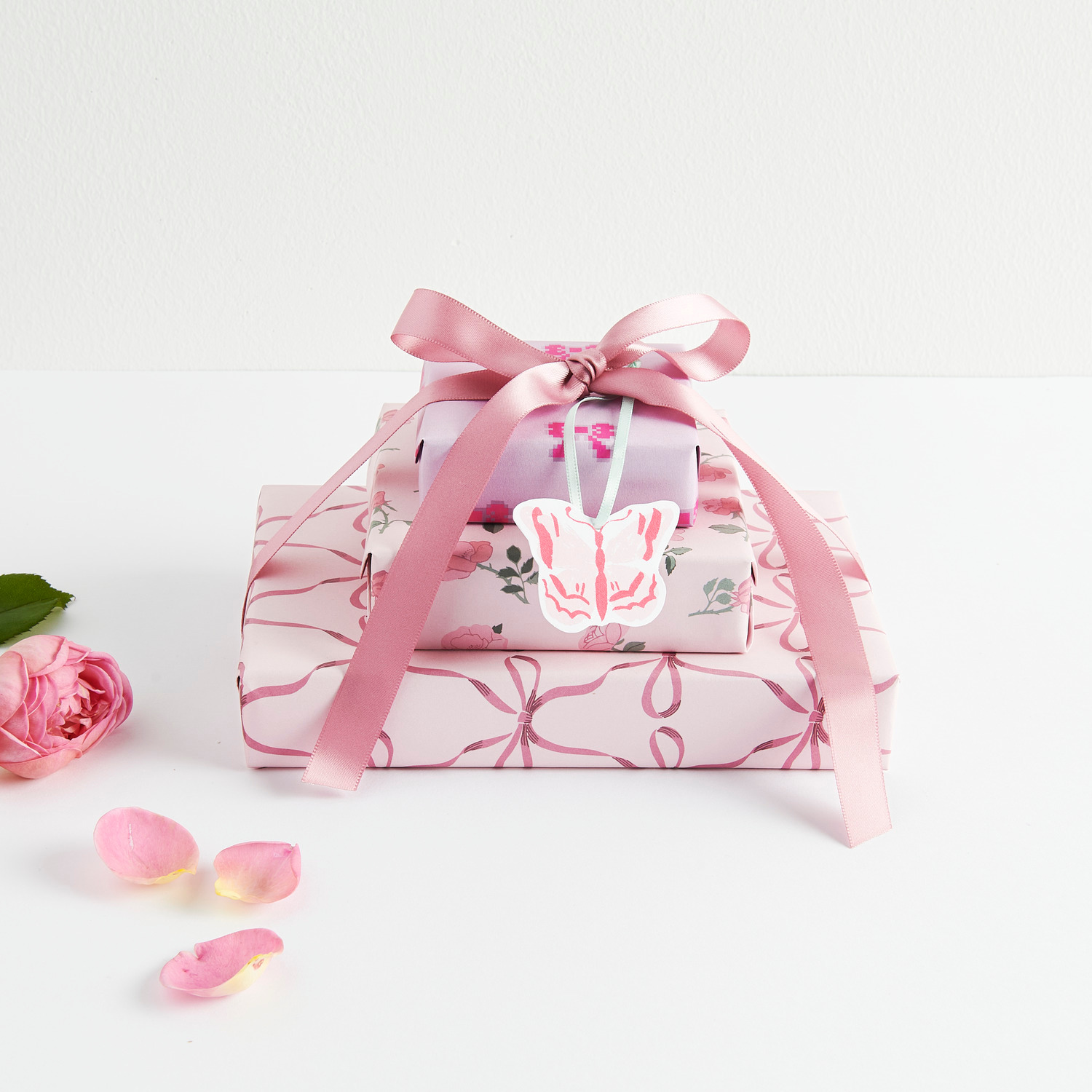 Paper Poetry Motivpapierblock La Vie En Rose: Zwei rosa Geschenke mit Schleifen und Schmetterlingsanhänger, dekoriert mit Rosenblättern.