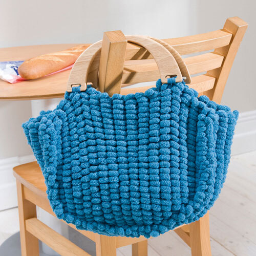 Pompon: Eine blaue, handgefertigte Tasche aus Rico Design Wolle, hängend an einem Holzstuhl.