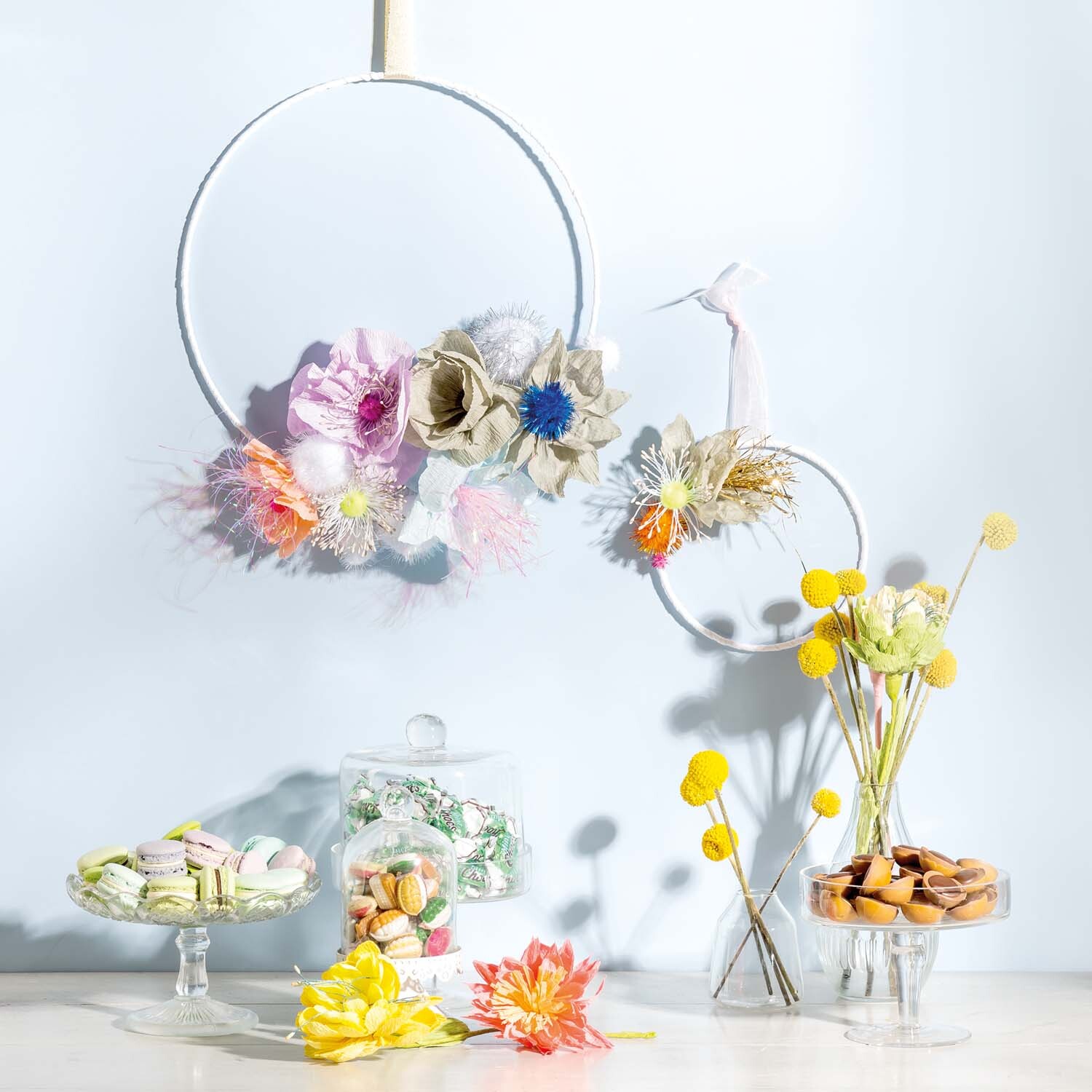 Bastelanleitung Blütenkranz: Ein dekorativer Kranz mit bunten Papierblüten, umgeben von Gläsern mit Süßigkeiten und gelben Blumen auf einem Tisch.