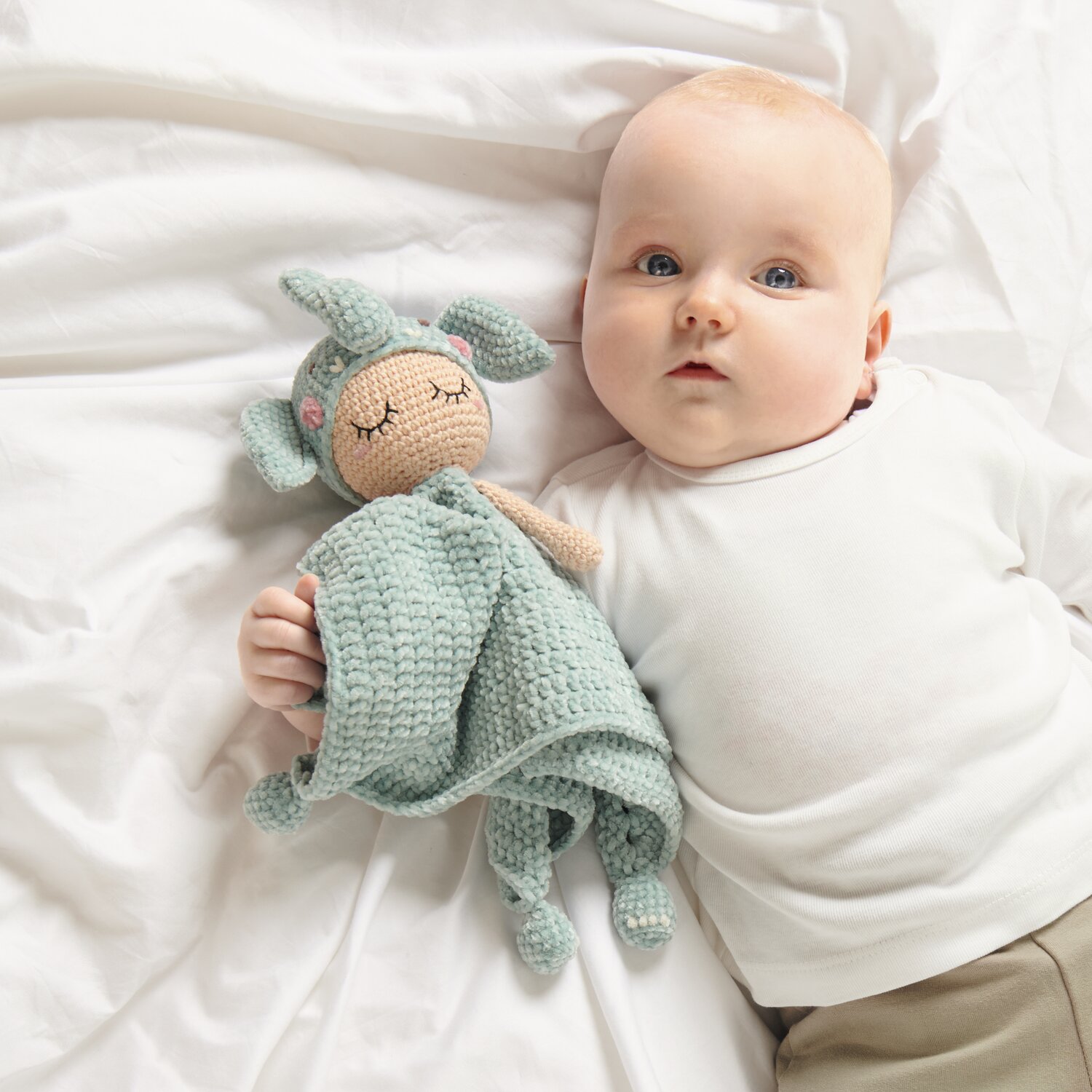 Ricorumi Baby Blankies Nilli Nilli: Ein Baby liegt auf weißer Bettwäsche mit einem gehäkelten Kuscheltier in Aqua.
