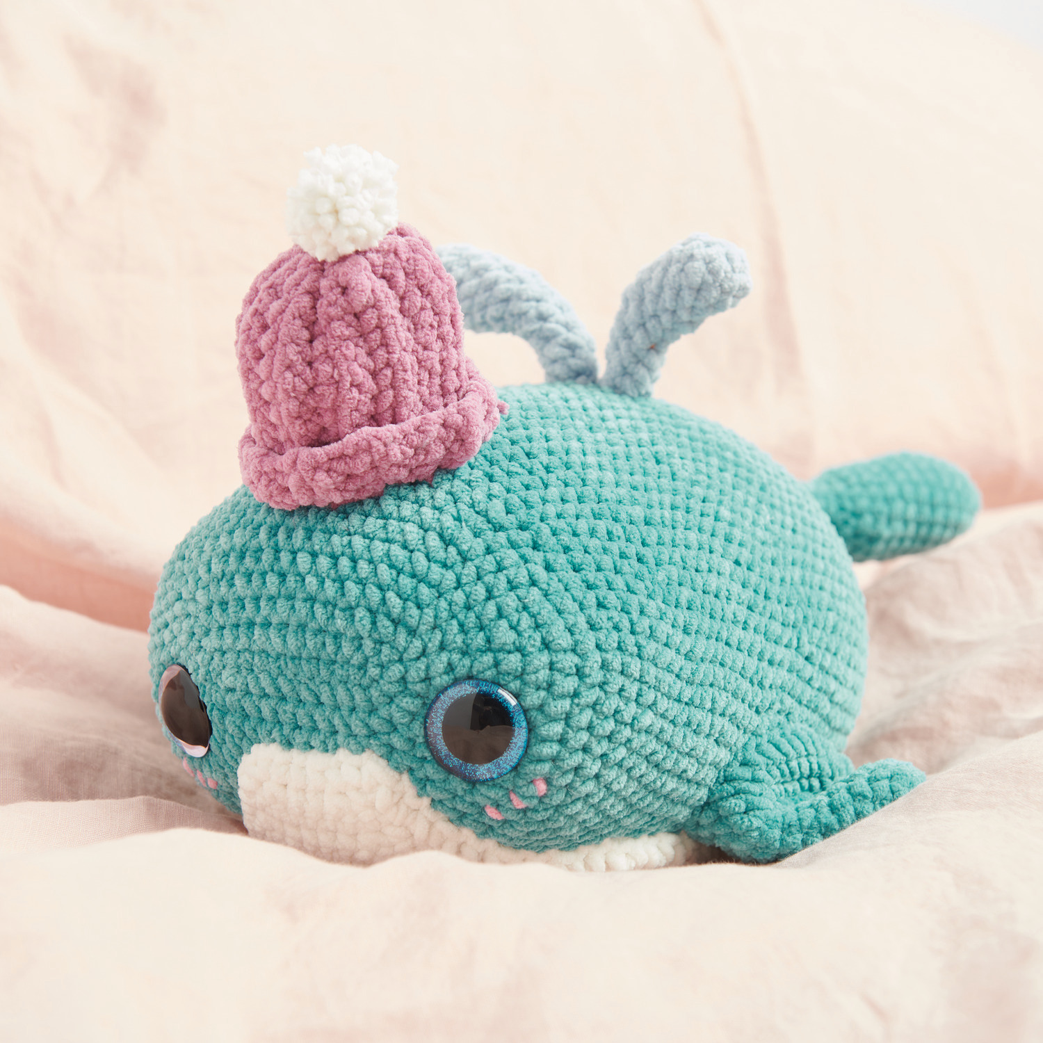 Häkelset Grandpa Whale Modell 06: Ein türkisfarbener Wal aus Chenille mit rosa Mütze auf beigem Hintergrund.
