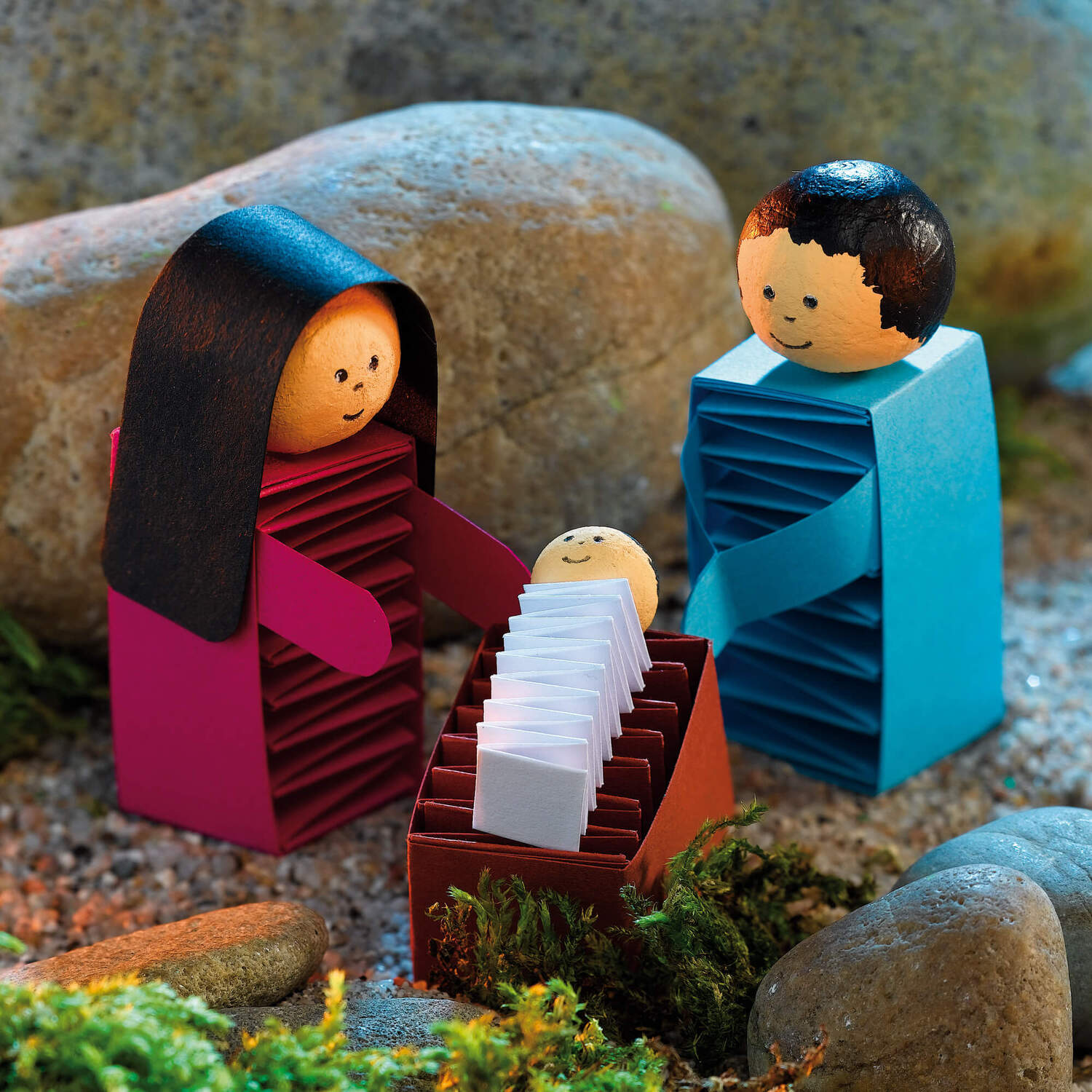 Bastelanleitung Weihnachtskrippe: Handgefertigte Figuren aus Papier in einer Krippenszene mit Steinen und Moos.