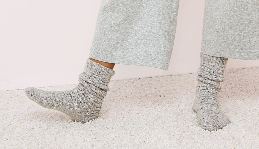Strickanleitung Wellnesssocken: Graue, gestrickte Socken auf Teppichboden, getragen von Person in grauer Hose.
