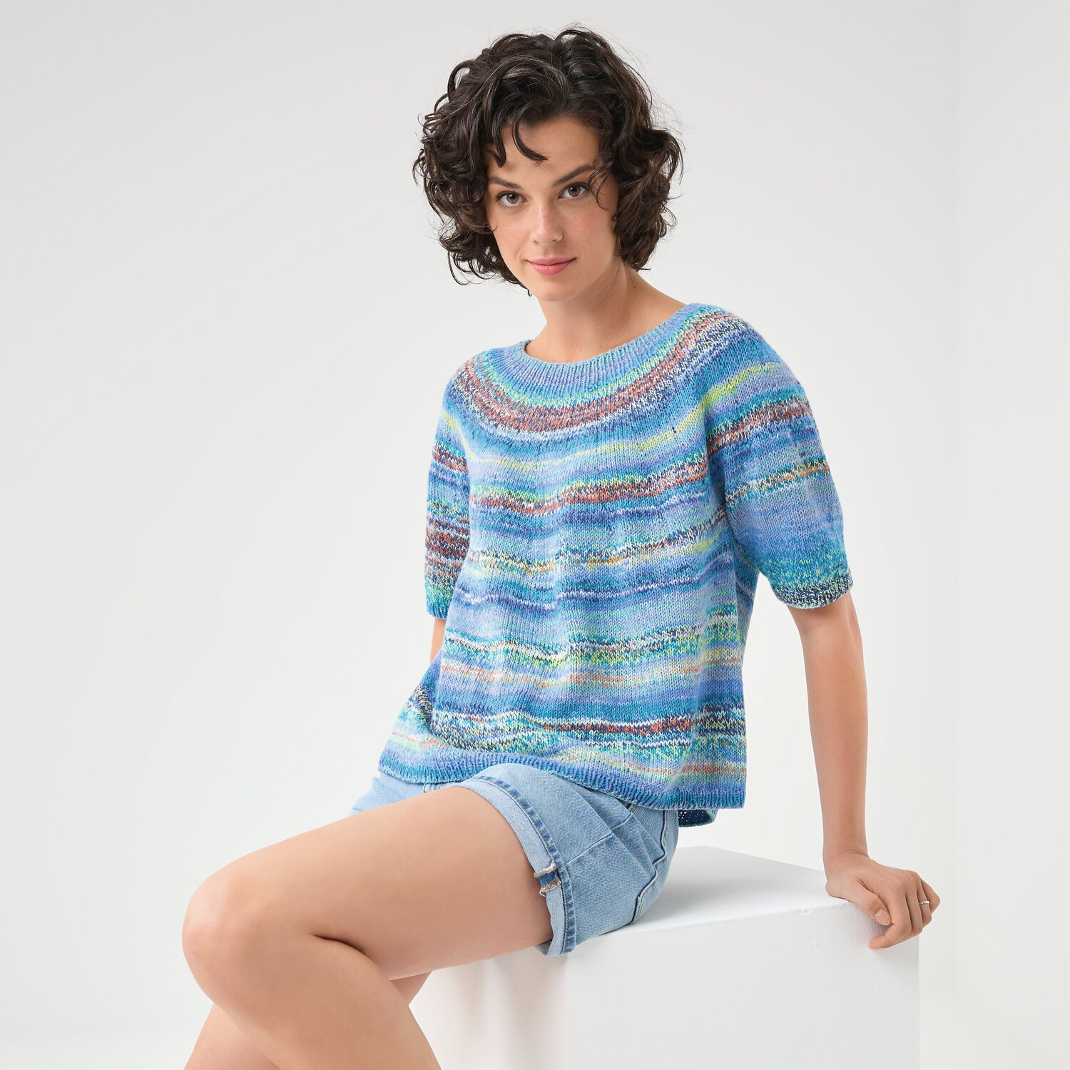 Strickset Shirt Modell 22: Frau in blauem, gestreiftem Shirt und Jeansshorts sitzt auf weißem Würfel.