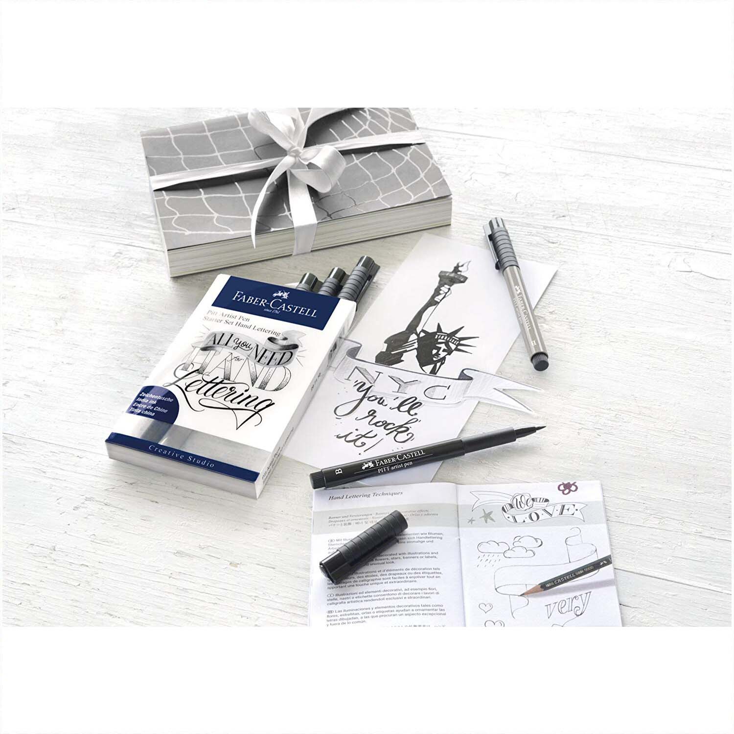 Tuschestift Pitt Artist Pen Handlettering Starter 9 Stück von Faber Castell auf einem Tisch mit Geschenkbox und Handlettering-Beispielen.