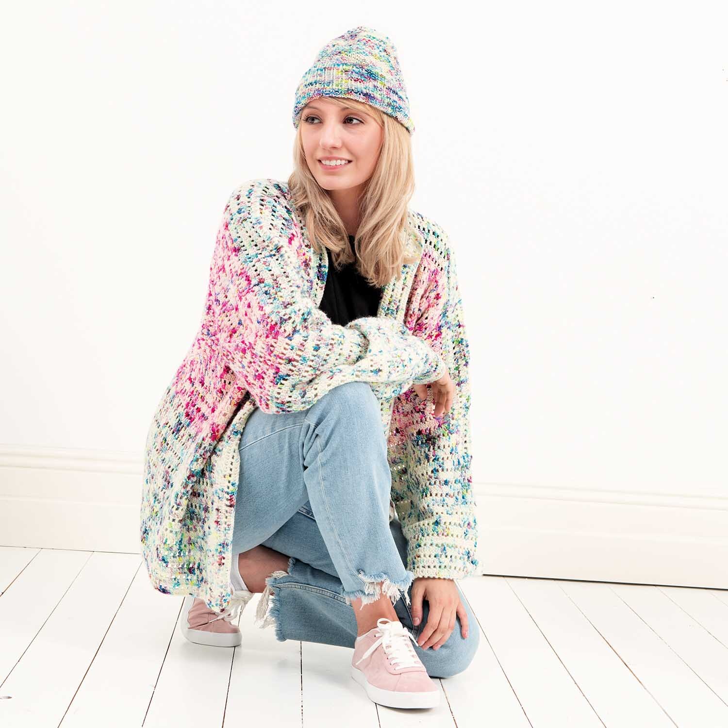 Luxury Hand-Dyed Happiness dk: Frau in gestrickter Jacke und Mütze in Natur-Pink von Rico Design, sitzt lächelnd auf weißem Boden.