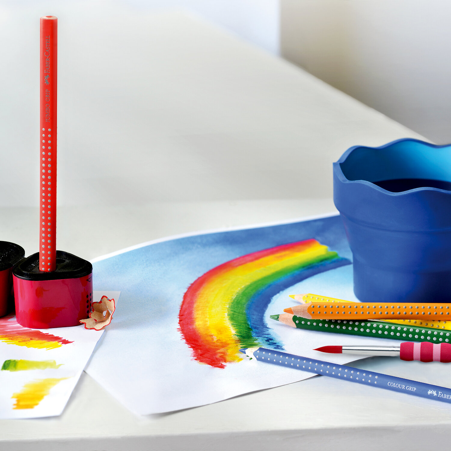 Colour Grip 2001: Bunte Faber Castell Stifte auf Tisch mit Regenbogenzeichnung und blauem Becher.
