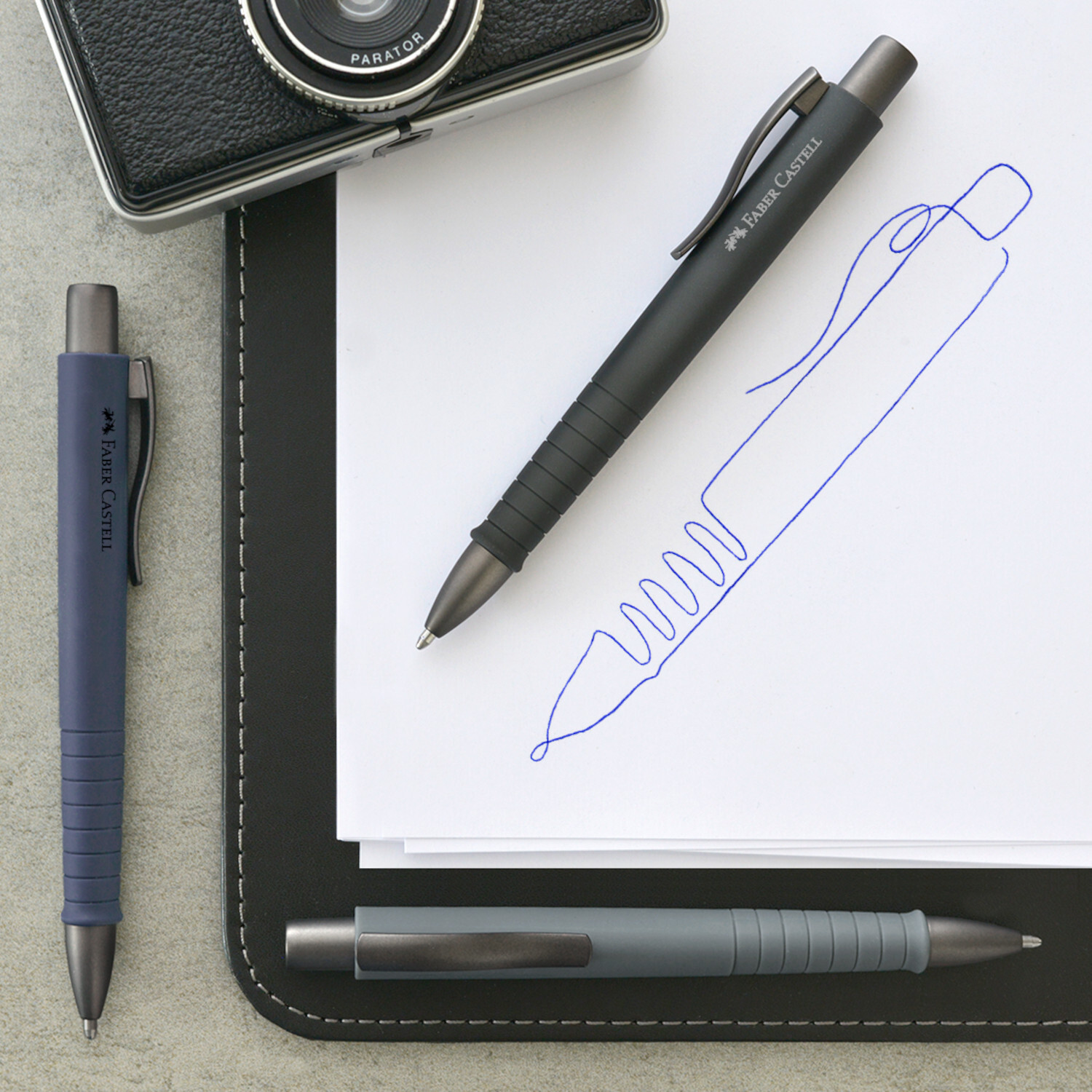 Kugelschreiber Poly Ball Urban XB von Faber Castell in Blau und Schwarz auf Notizblock neben Kamera.