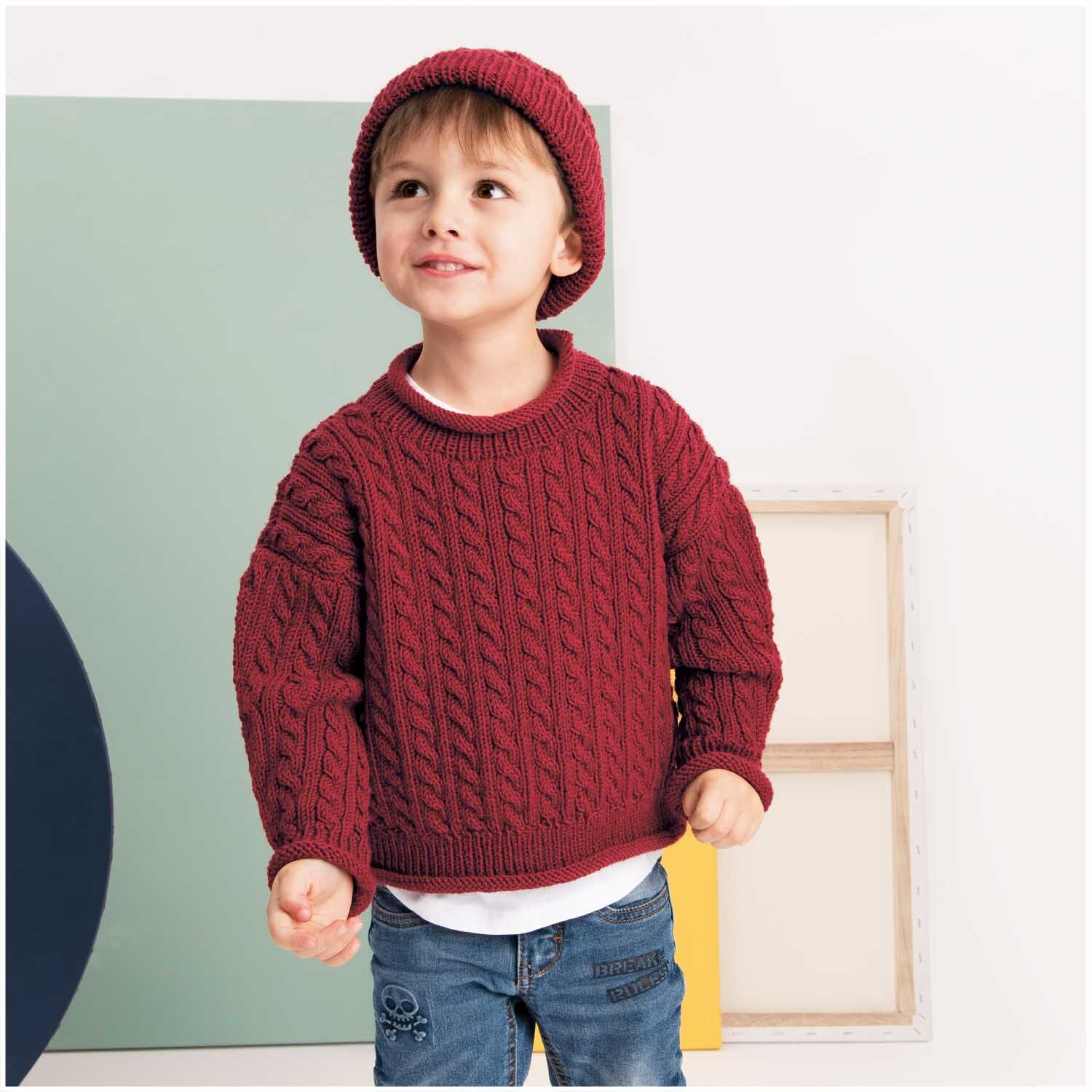 Essentials Merino dk: Kind trägt roten Strickpullover und Mütze von Rico Design, lächelnd.