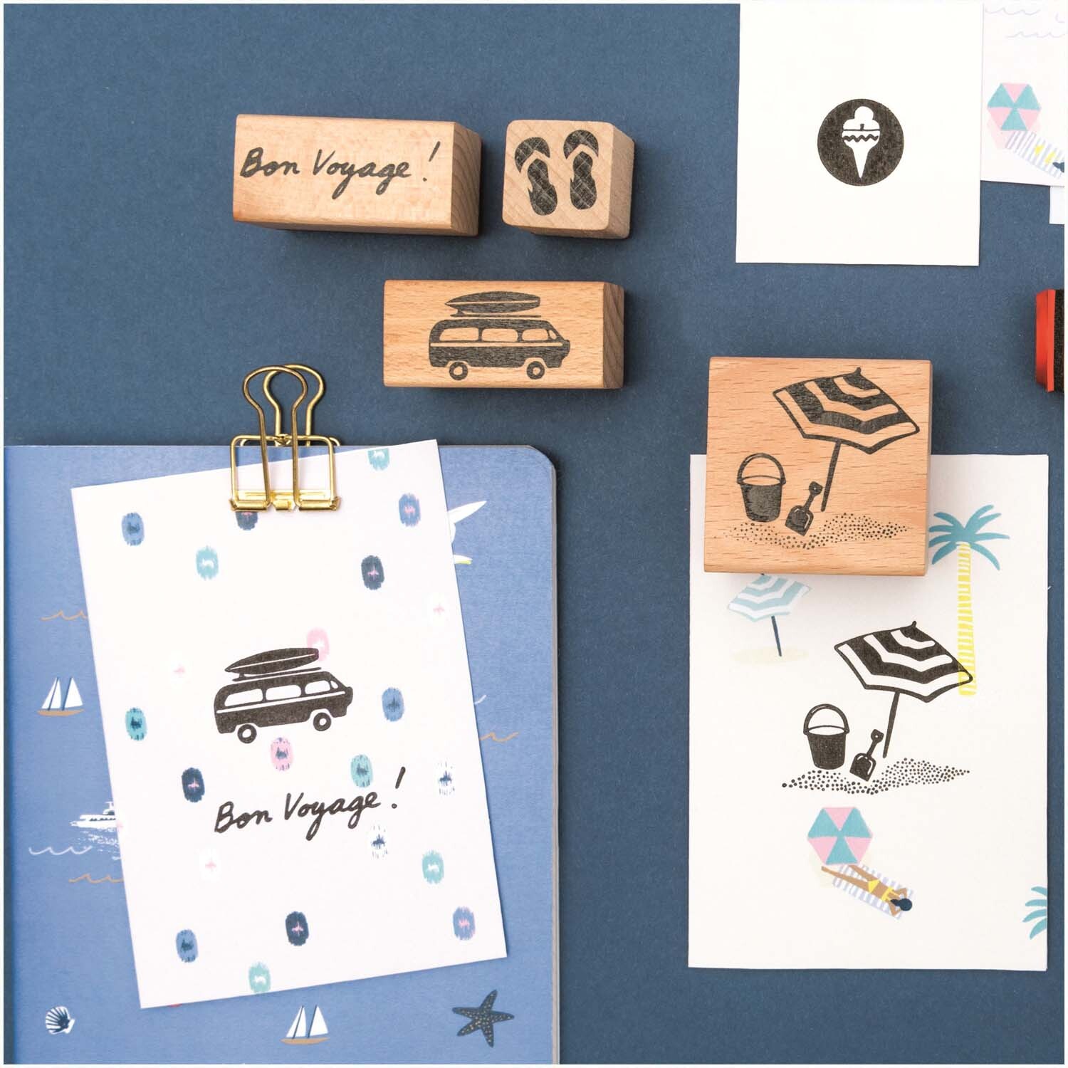 Paper Poetry Stempelset Bon Voyage: 7-teiliges Set auf blauem Hintergrund mit Reise- und Strandmotiven von Rico Design.