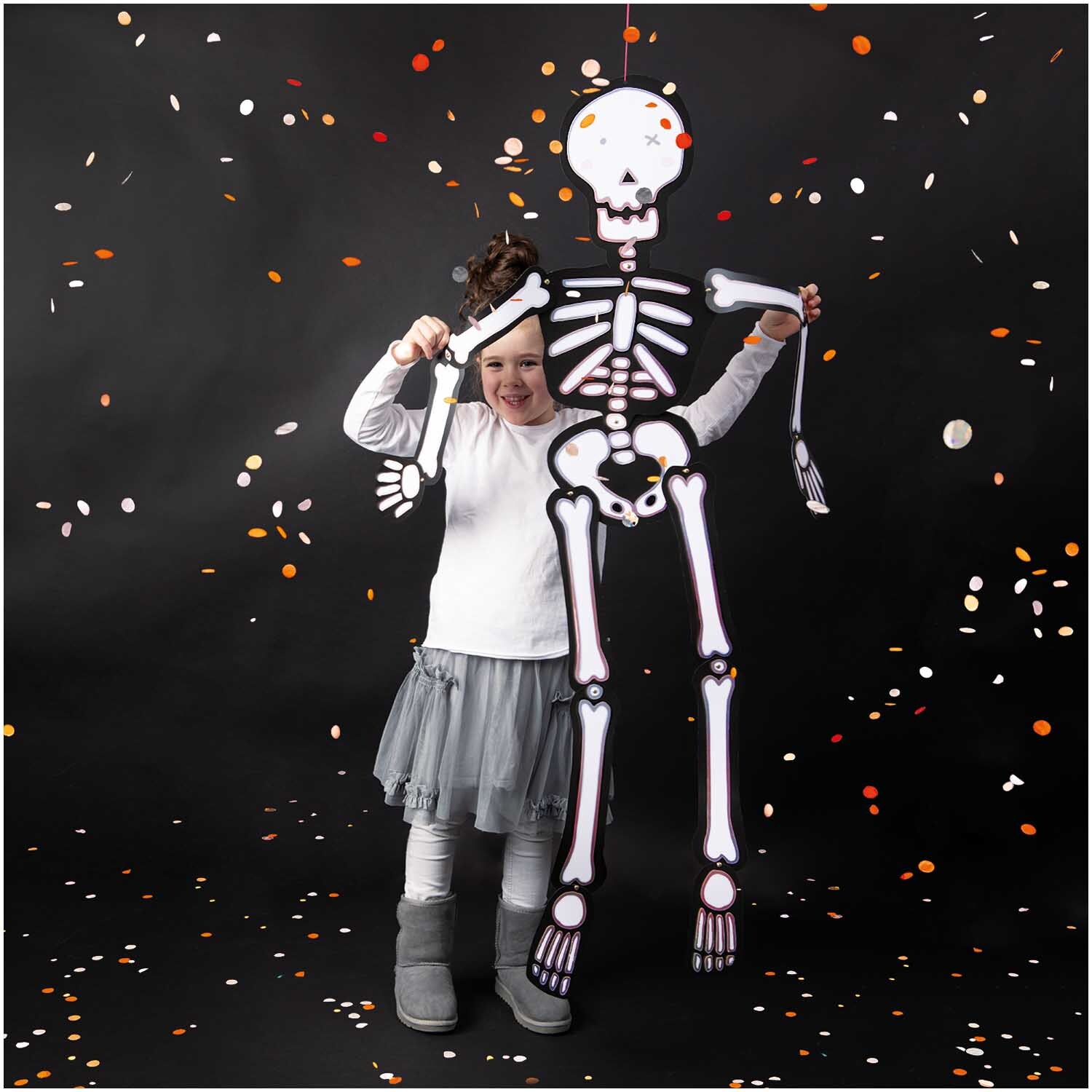 Paper Poetry Hänge-Skelett groß 32x140cm von Rico Design: Kind hält ein großes Skelett vor schwarzem Hintergrund mit bunten Konfetti.