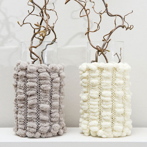 Pompon: Zwei Glasvasen mit gestrickten Überzügen in Grau und Weiß, dekoriert mit Zweigen.