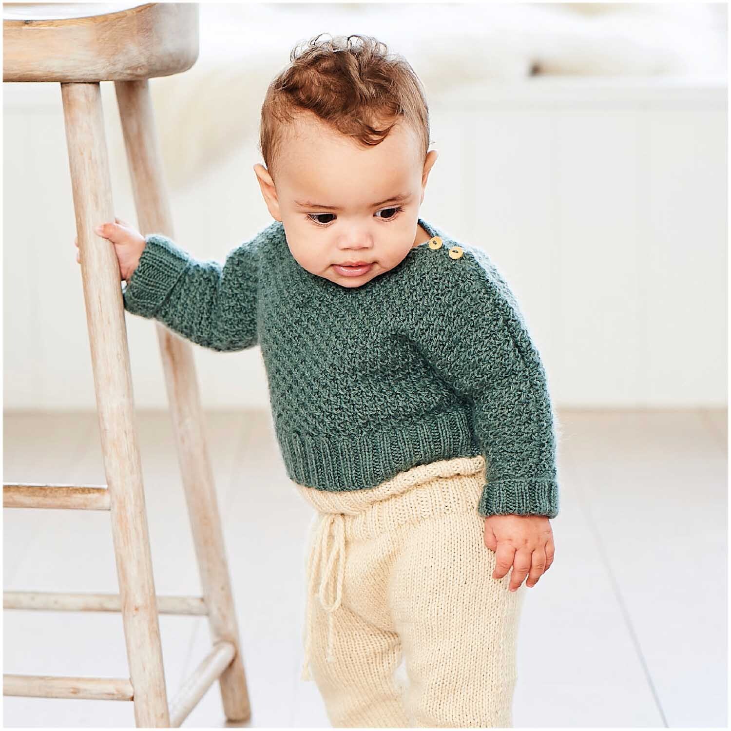 Rico Baby Dream dk uni - A Luxury Touch: Kleinkind in cremefarbener Hose und grünem Strickpullover.