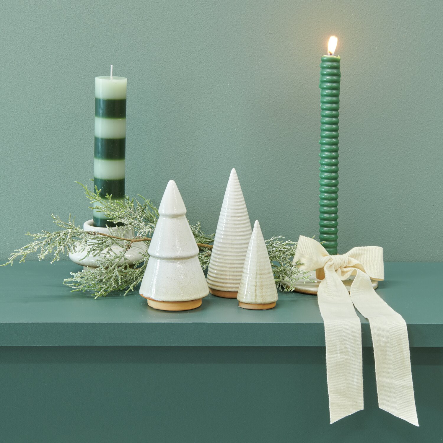 Porzellan Tannenbaum Geriffelt Beige neben gestreifter Stabkerze und Ringkerze auf grünem Regal, dekoriert mit Baumwollband und Zweigen.