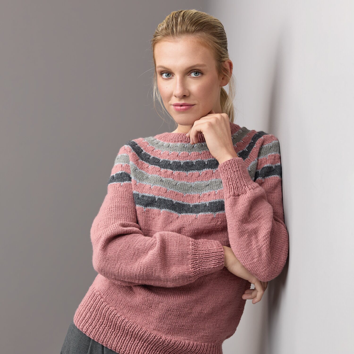 Jubiläums-Strickset Pullover Modell 08: Frau trägt gestrickten Pullover mit Streifenmuster in Rosa und Grau, lehnt an einer Wand.