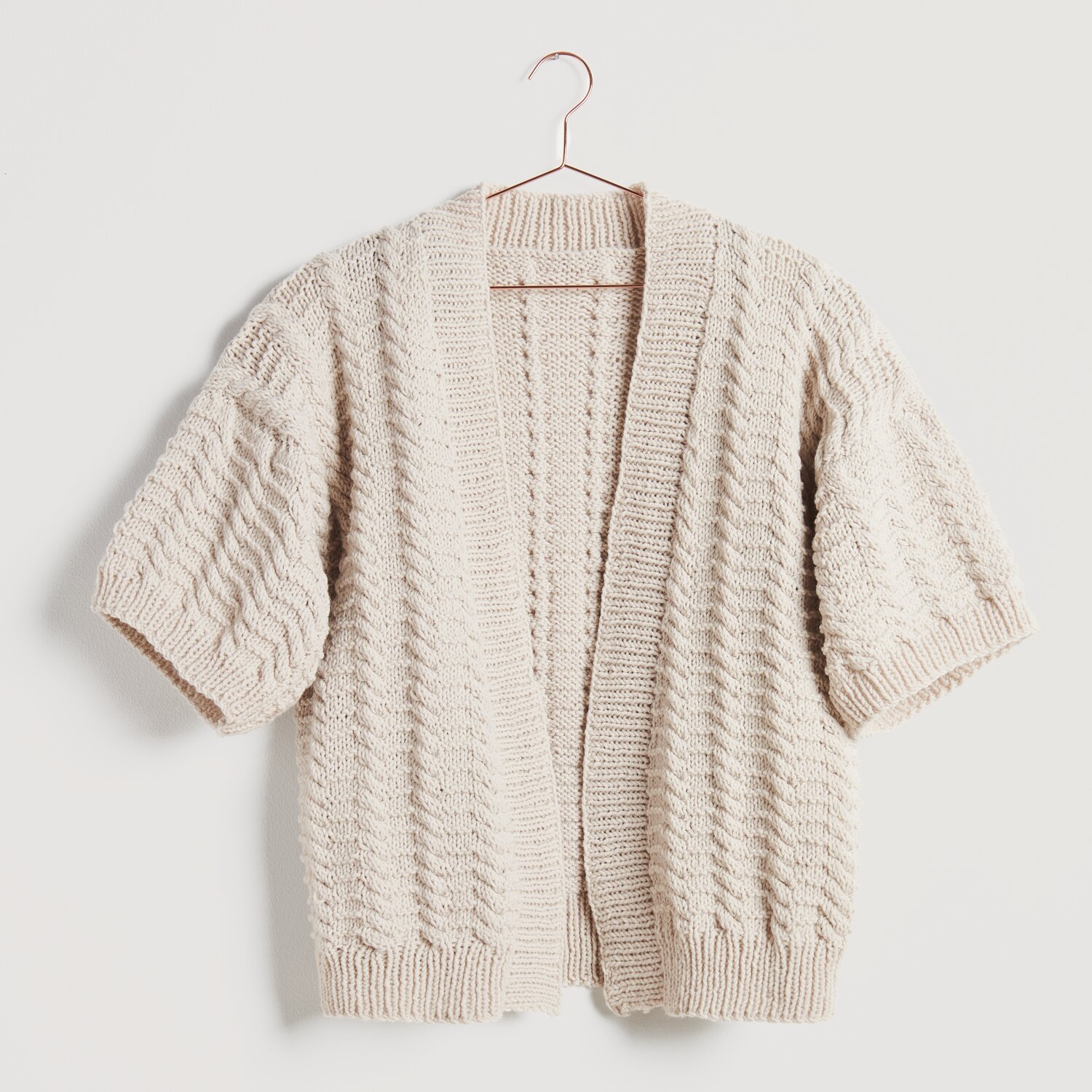 Strickset Cardigan Modell 03: Beigefarbener, kurzärmeliger Cardigan mit Zopfmuster auf einem Kleiderbügel.