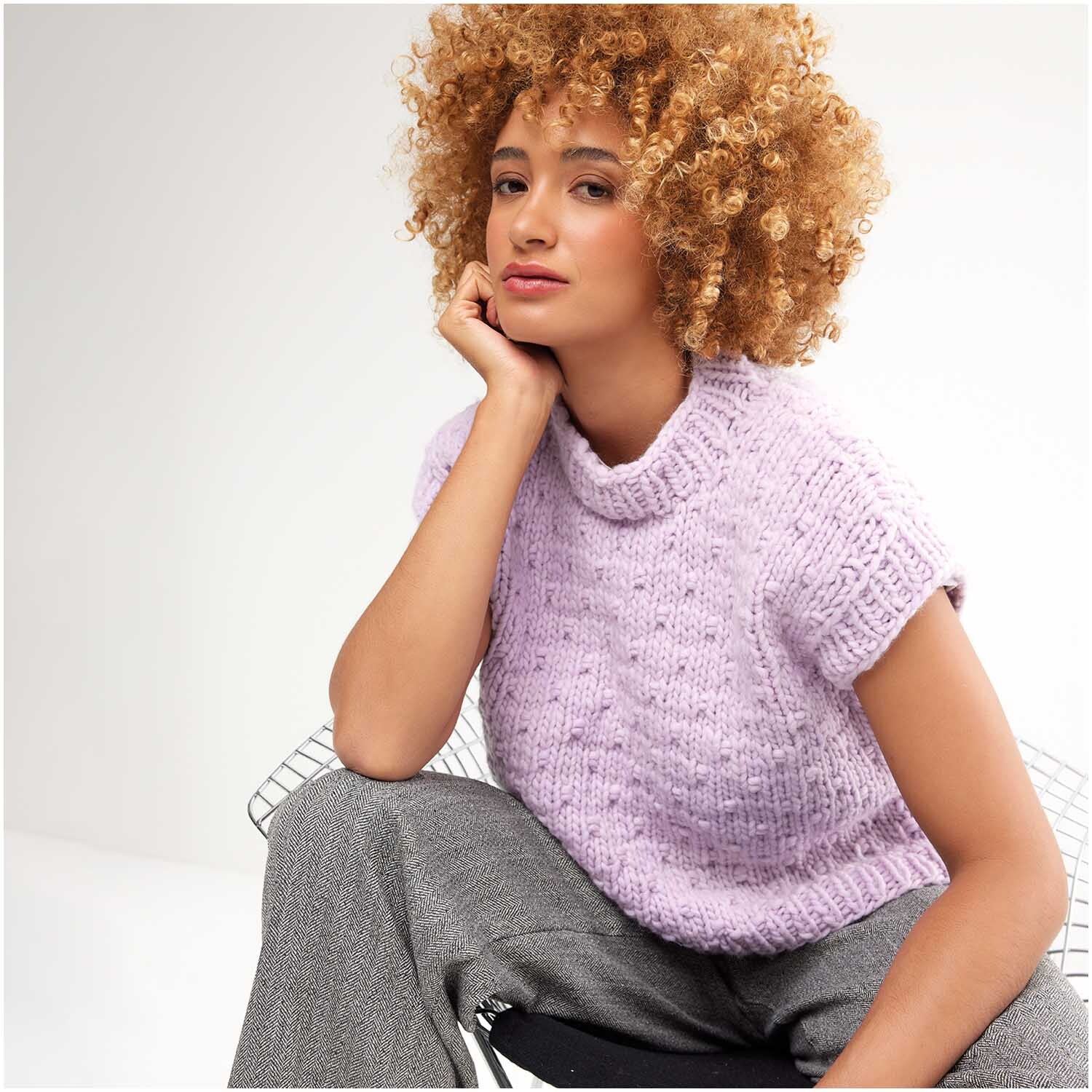 Strickset Pullunder Modell 02: Frau trägt lila Pullunder, sitzt entspannt, lockiges Haar, graue Hose.