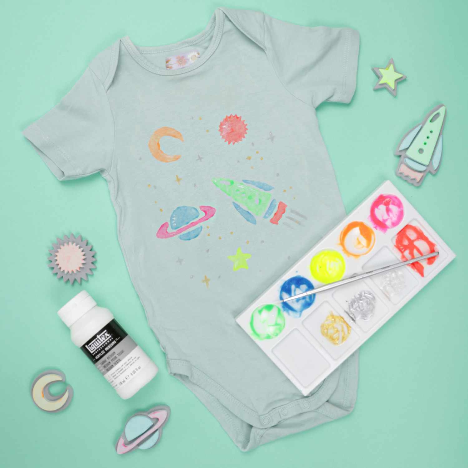 Anleitung Babybody mit bunten Weltraummotiven, Acrylfarbe und Stempeln auf mintgrünem Hintergrund.