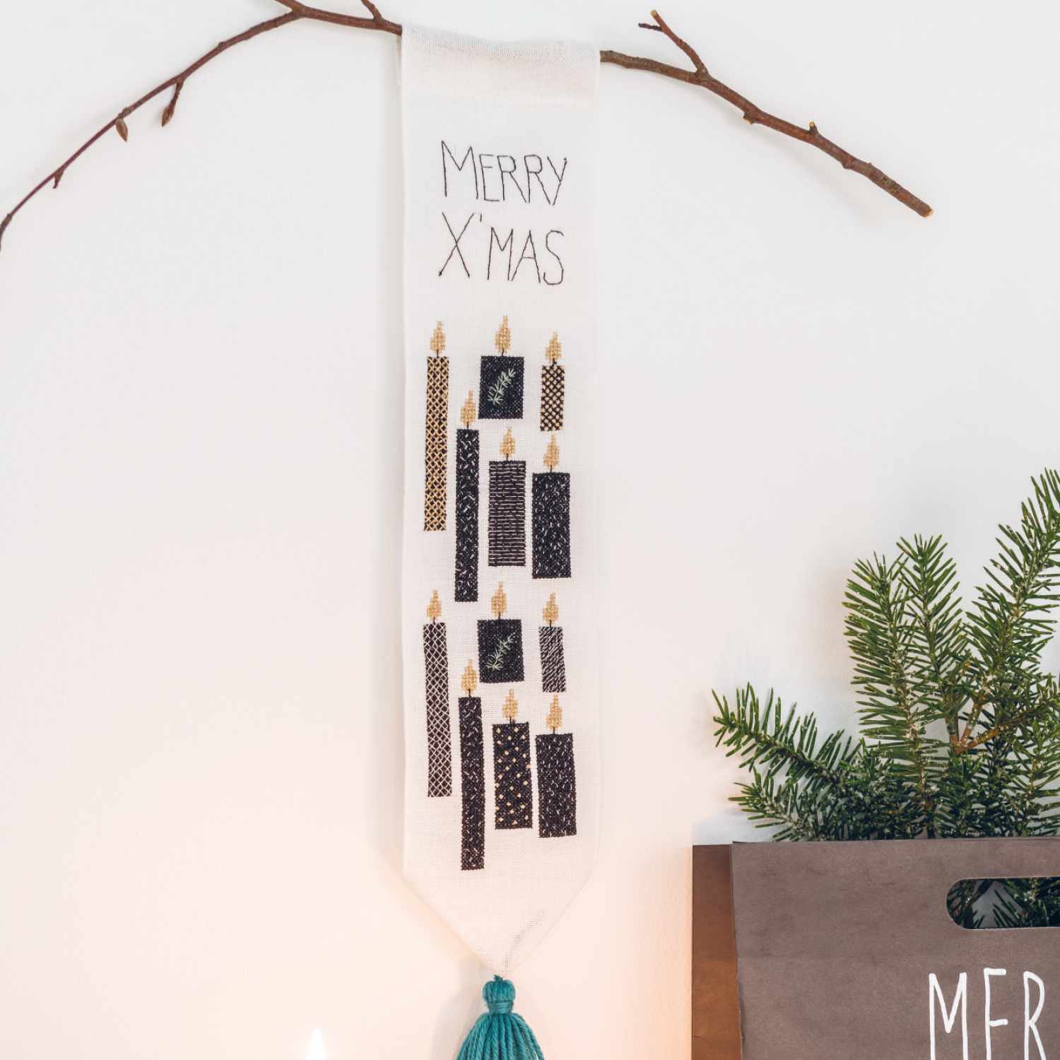 Stickanleitung Weihnachtlichen Wandbehang gestalten: Weißer Wandbehang mit schwarzen Kerzen und grüner Quaste, dekoriert mit Tannenzweigen.