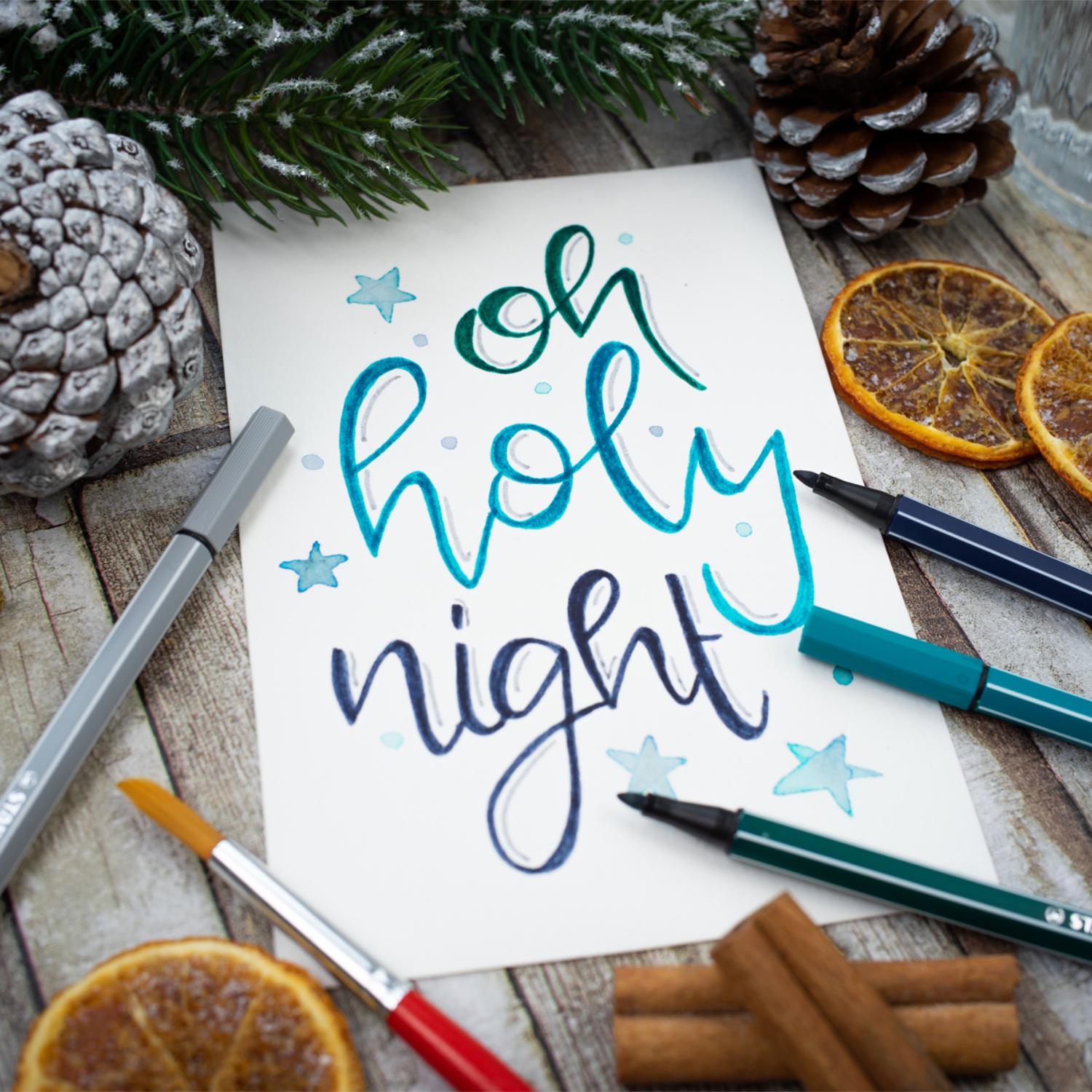 Pen 68: Handgeschriebener Text "oh holy night" auf Papier, umgeben von Stiften, Tannenzapfen und getrockneten Orangenscheiben.