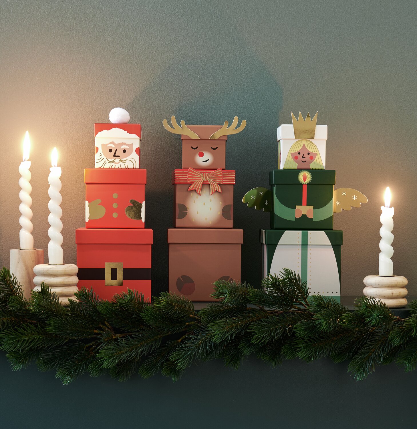 Geschenkboxen-Set Weihnachtsmann: Vier dekorative Boxen in Rot, Braun und Grün, umgeben von Kerzen und Tannenzweigen.