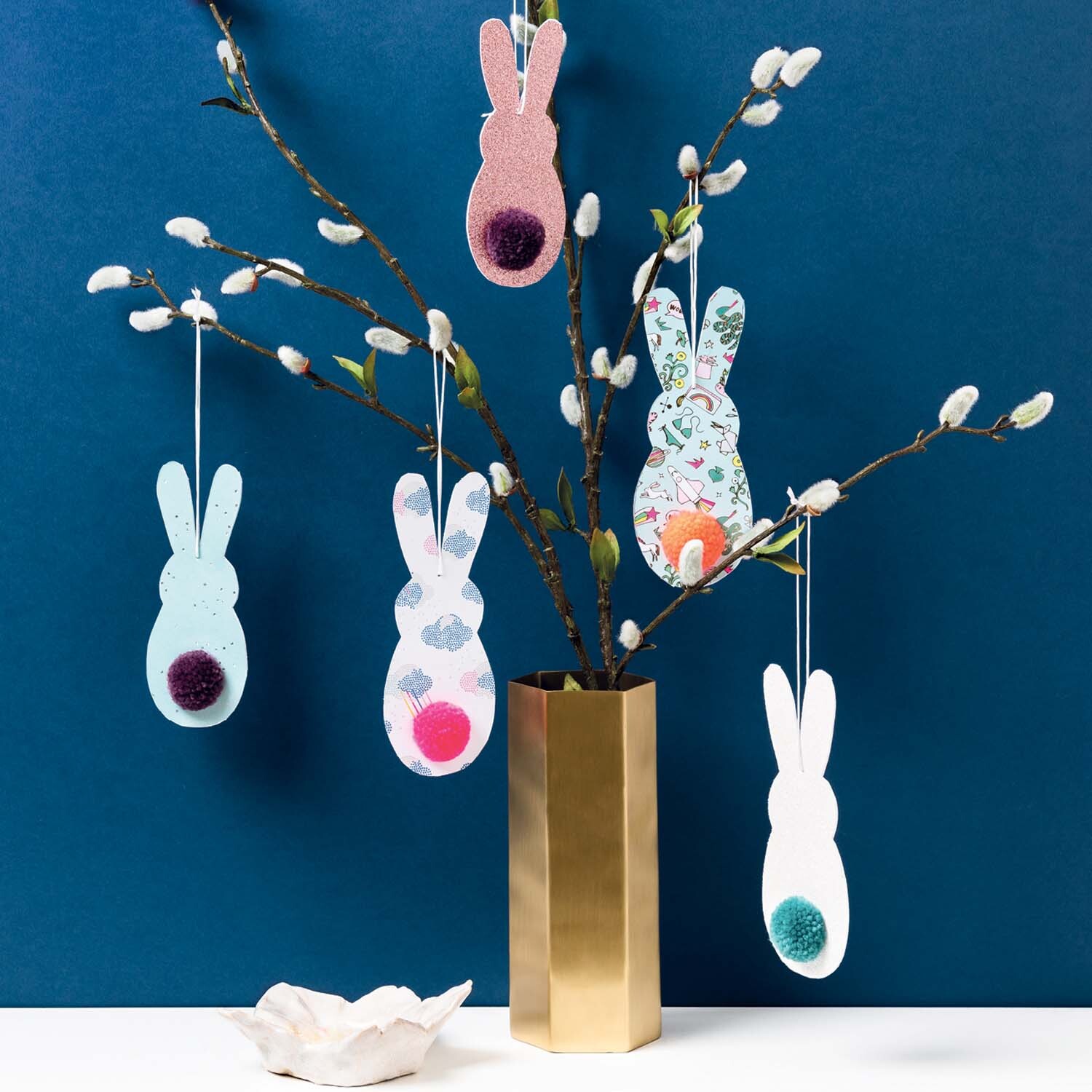 Bastelanleitung Hasenanhänger: Fünf bunte Hasenanhänger an Zweigen in einer goldenen Vase vor blauem Hintergrund.