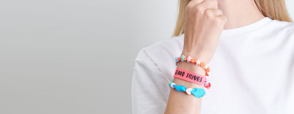 Armgelenk mit einem orangen Armband mit bunten Sternen, ein pinkes Armband mit schwarzem Schriftzug und einem blauen Armband mit Herz und runder Perle.