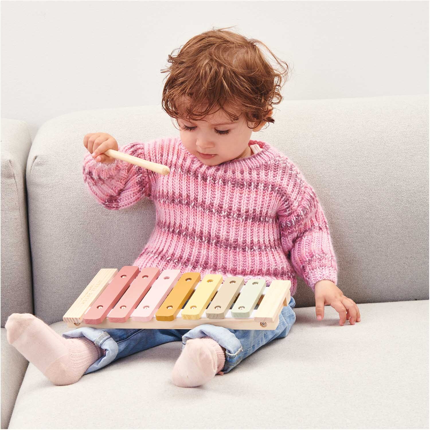 Rico Baby Dream dk A Luxury Touch: Ein Kind im rosa Pullover spielt auf einem bunten Xylophon auf dem Sofa.