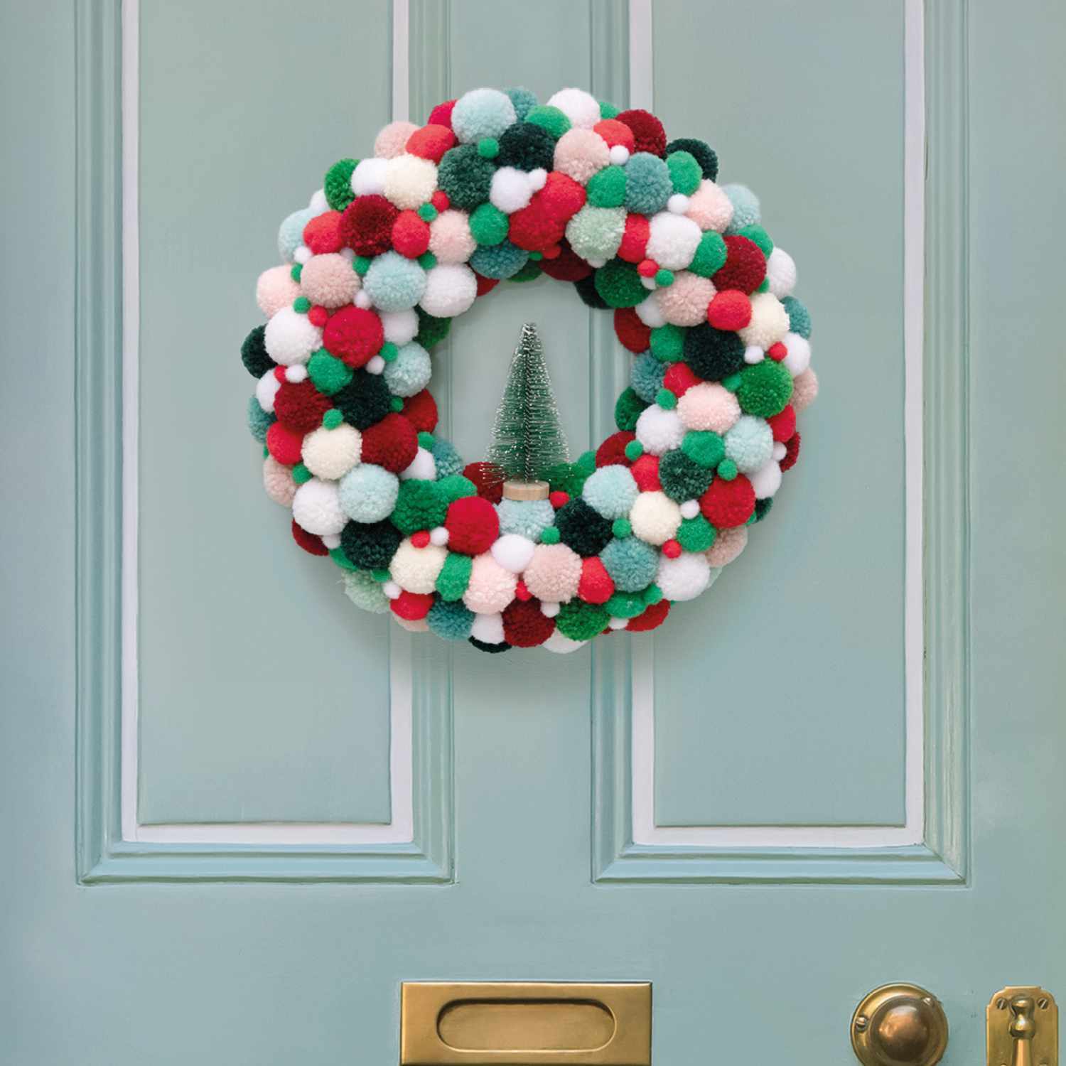 Bastelanleitung weihnachtlicher Pompon-Türkranz: Bunter Kranz aus Pompons an einer blauen Tür mit goldener Türklinke.