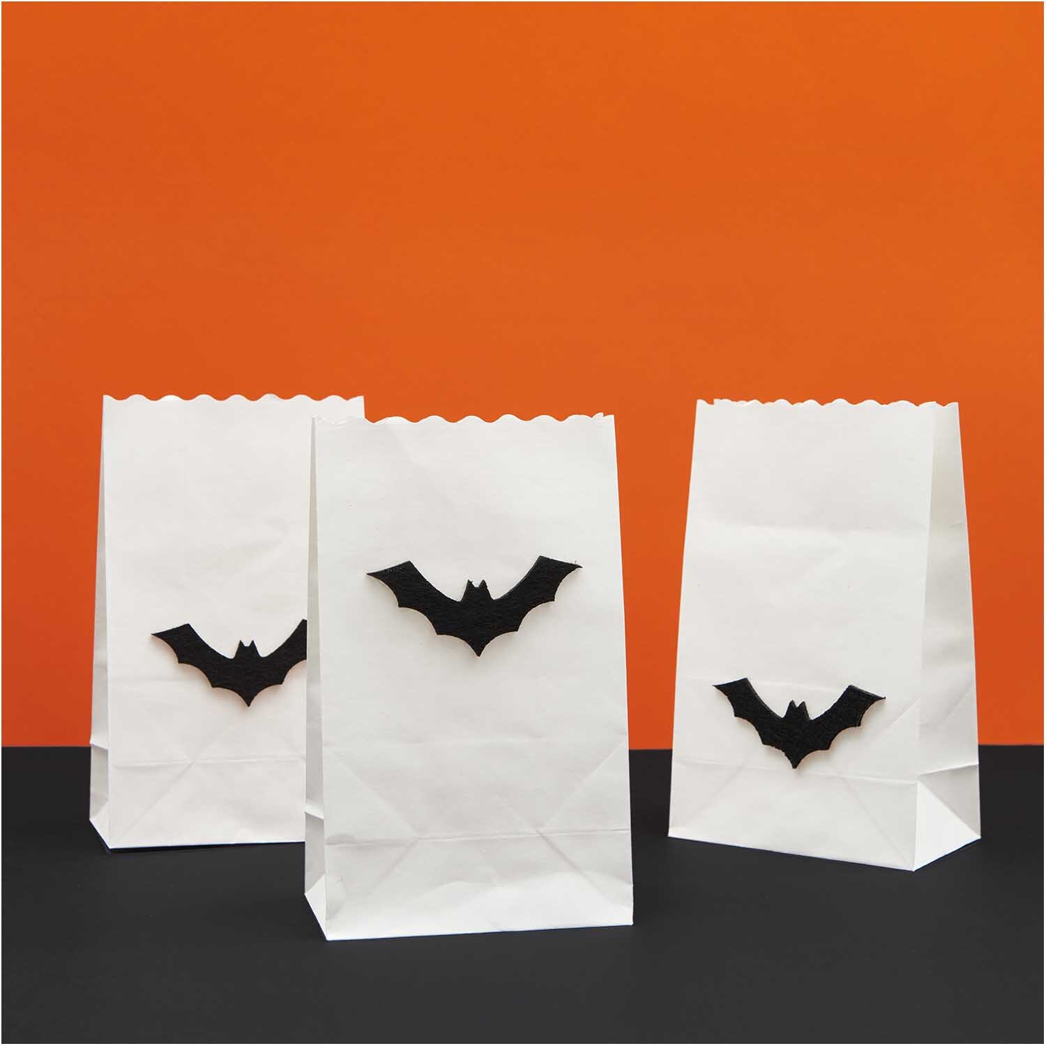 Filzstreu Fledermaus schwarz 6 Stück: Weiße Papiertüten mit schwarzen Fledermausmotiven vor orangefarbenem Hintergrund.